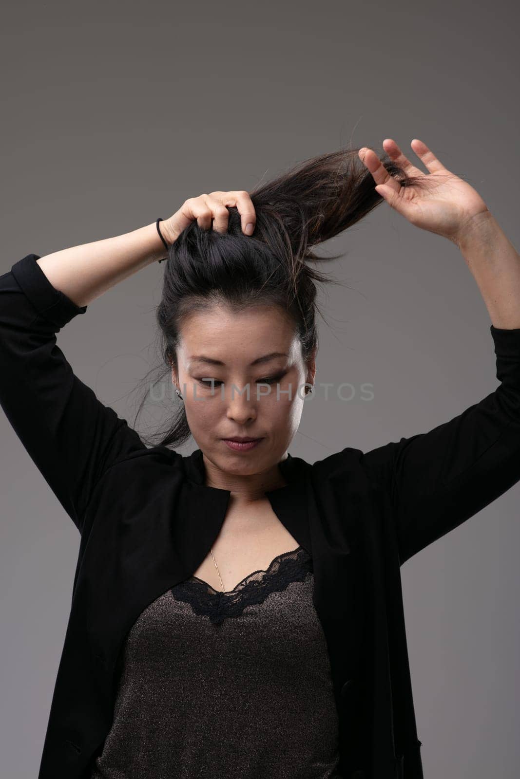 Attractive stylish Asian woman playing with her long hair holding it up in a ponytail above her head over a grey studio background