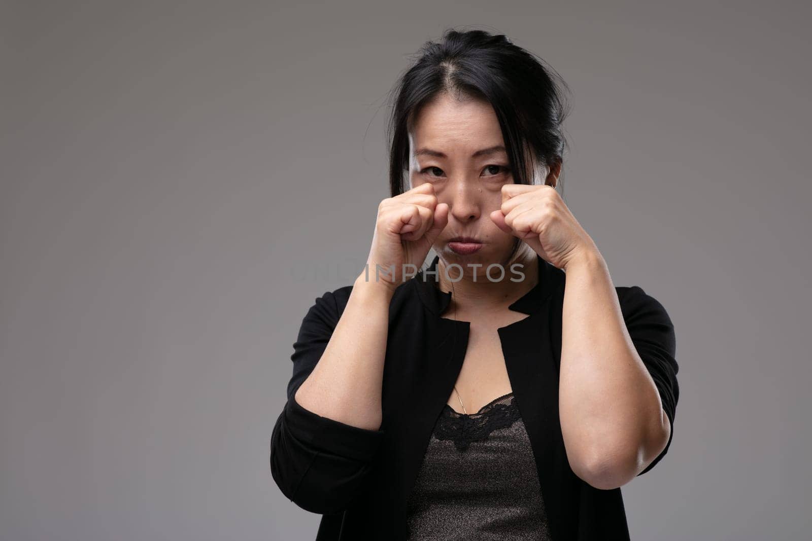 Upset morose Asian woman standing crying raising her hands to her eyes in sorrow over a grey studio background with copyspace
