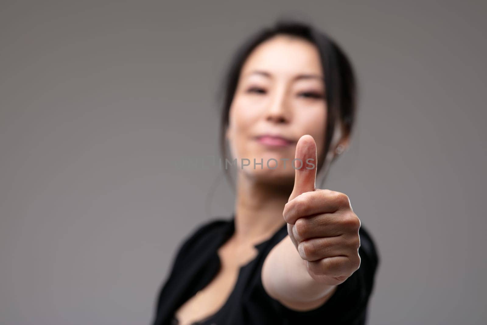 Elated triumphant Asian woman giving a thumbs up gesture of success and approval with focus to her hand over a grey studio background with copyspace