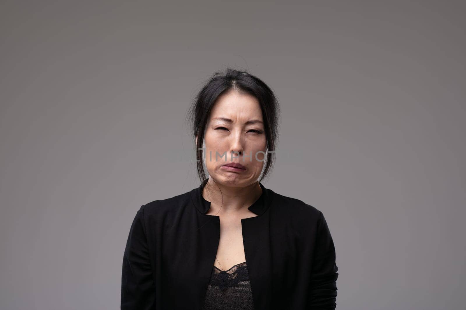 Tearful sorrowful Asian woman grimacing and screwing up her face in a frontal view over a grey studio background with copyspace