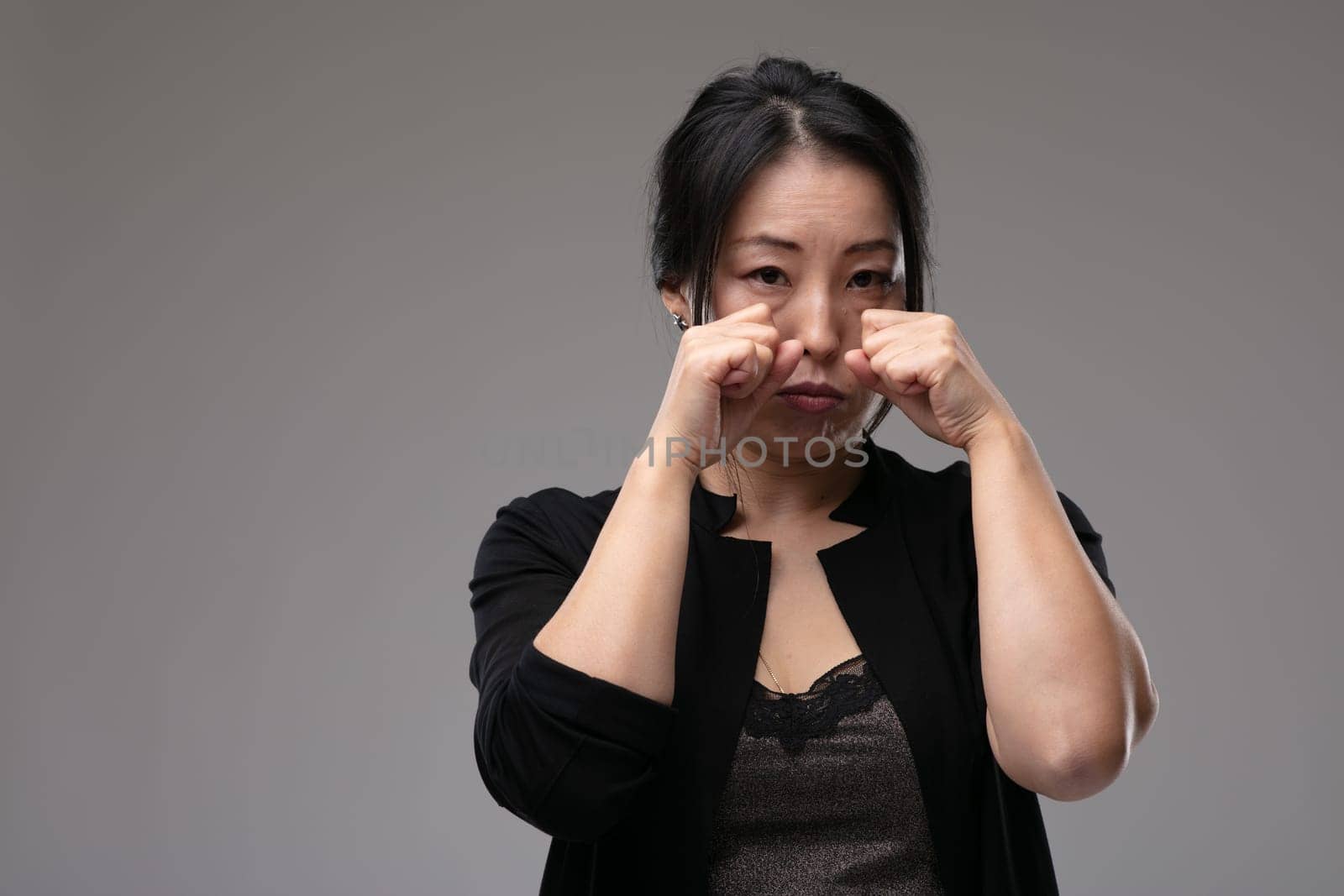 Sorrowful sad Asian woman crying raising her hands to her tearful face over a grey studio background with copyspace