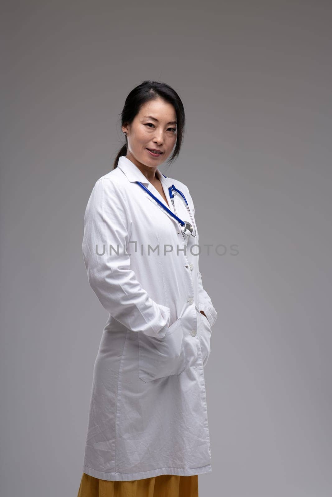 Thoughtful female Asian doctor or nurse in lab coat standing sideways looking t the camera with a quiet friendly smile over a grey studio background with copyspace