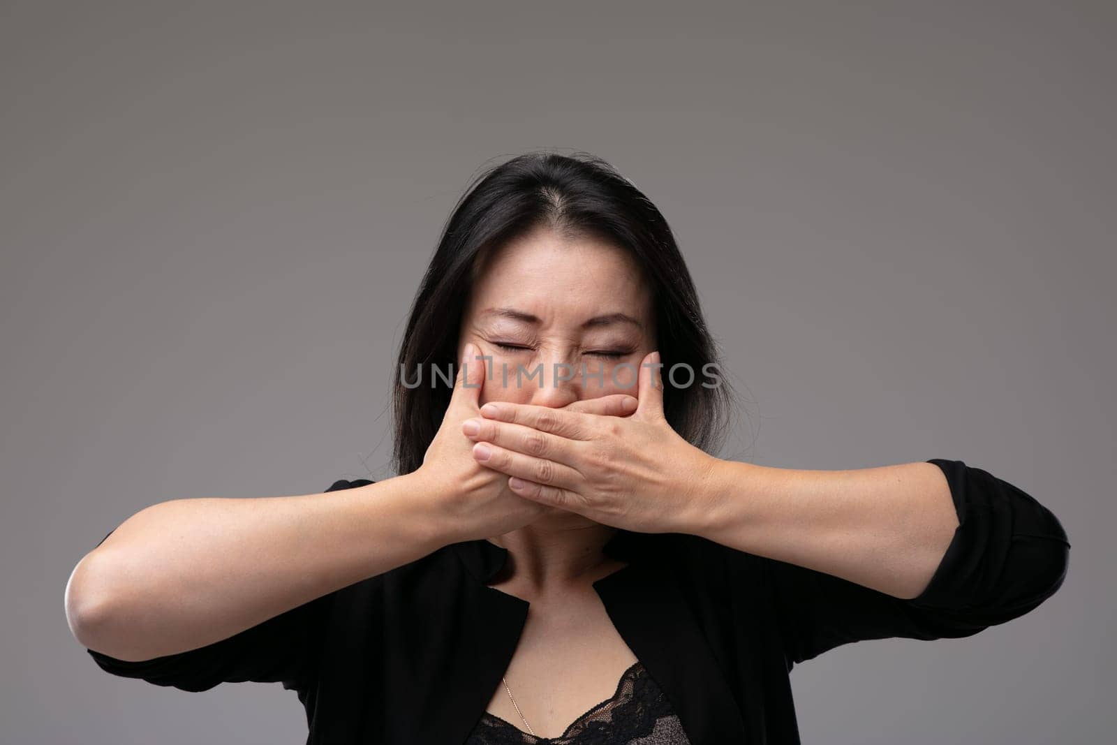 Asian woman with closed eyes covering her mouth with her hands in a concept of the metaphor see no evil, hear no evil speak no evil, over a grey studio background