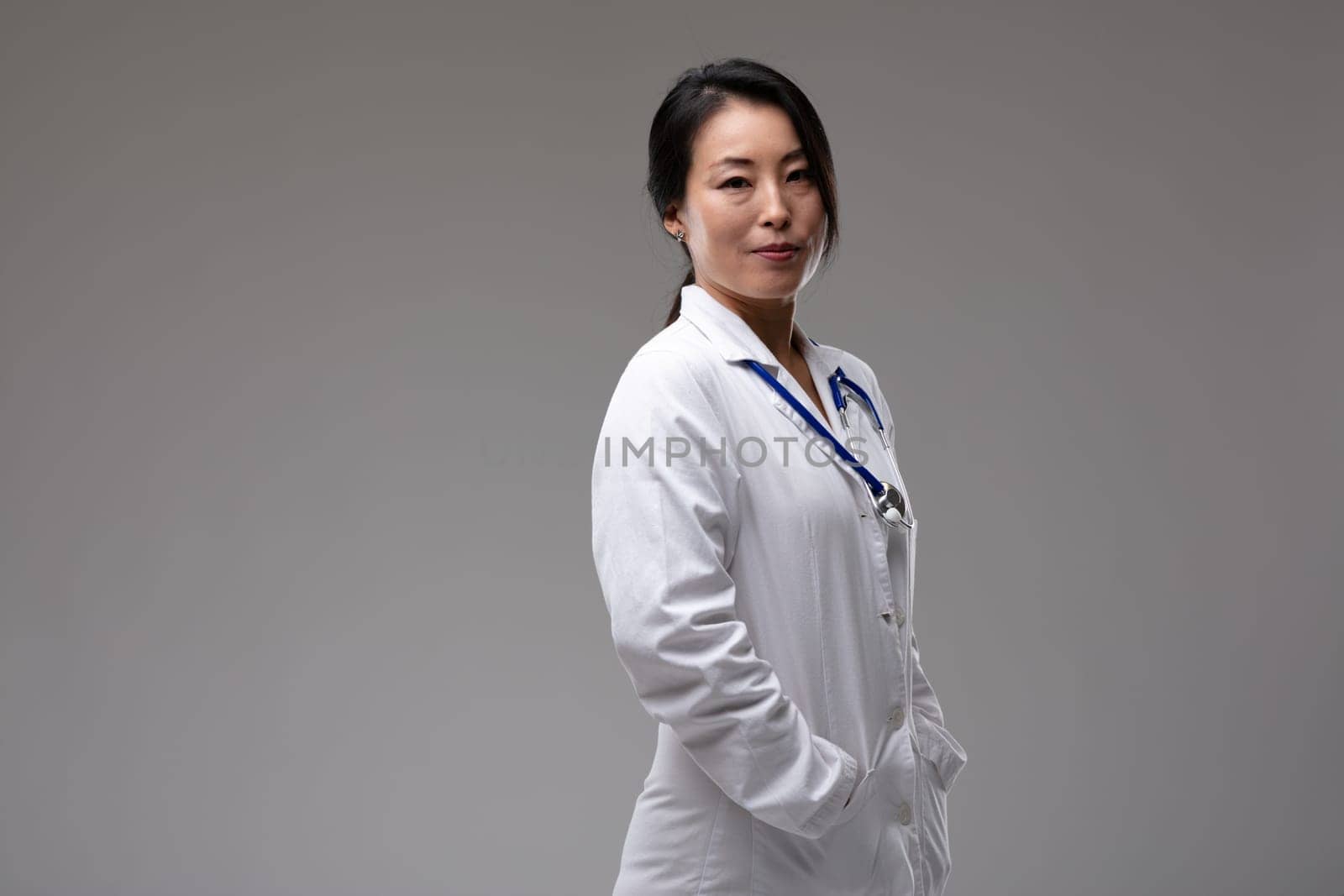 Thoughtful confident Asian woman doctor or nurse in lab coat with stethoscope turning to look at the camera with a quiet smile over grey with copy space