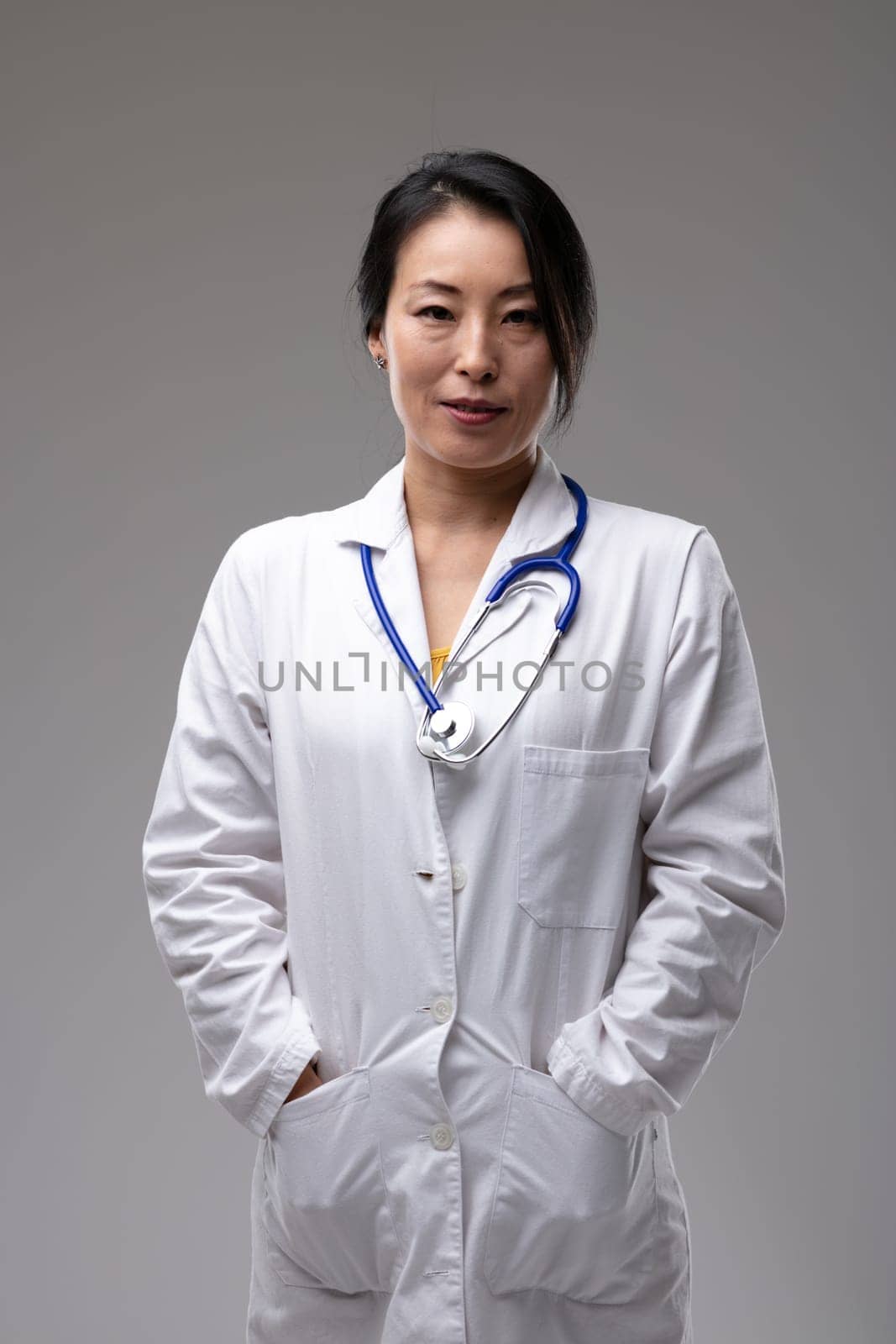 Confident relaxed Asian woman doctor with her hands in pockets in the pockets of her lab coat and stethoscope around her neck isolated on grey