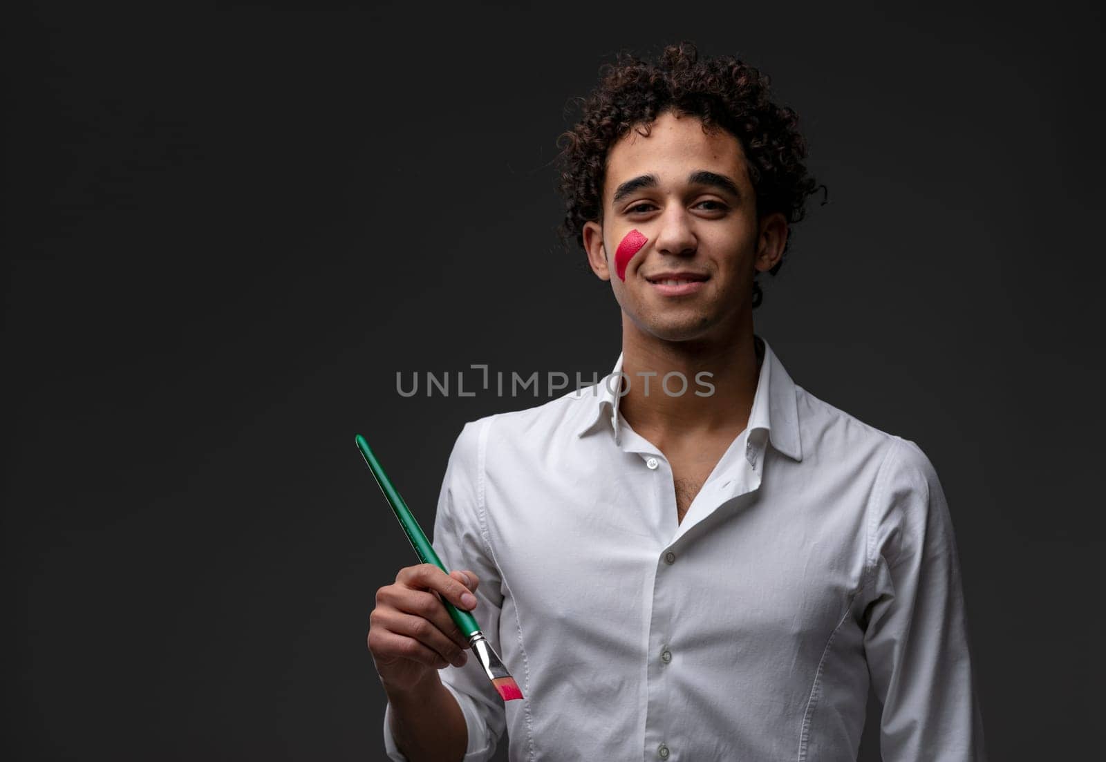 Smiling young curly haired artist holding a paintbrush with a colorful daub of red paint on his cheek as he smiles quietly at the camera over a dark background