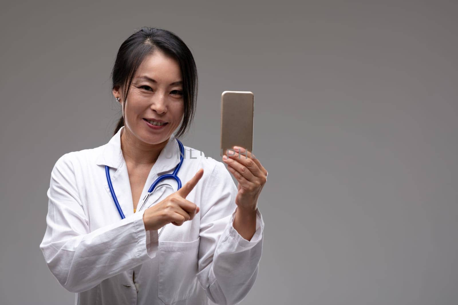 Smiling female Asian doctor or nurse pointing to her mobile phone isolated on grey with copy space