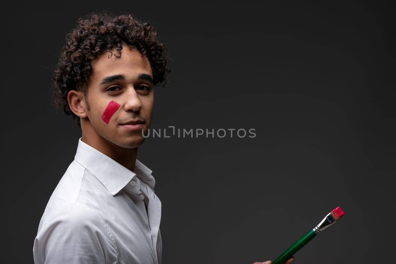 Serious thoughtful young male artist with red paint on cheek turning to look at the camera as he holds a paintbrush in his hand with copyspace over a dark studio background