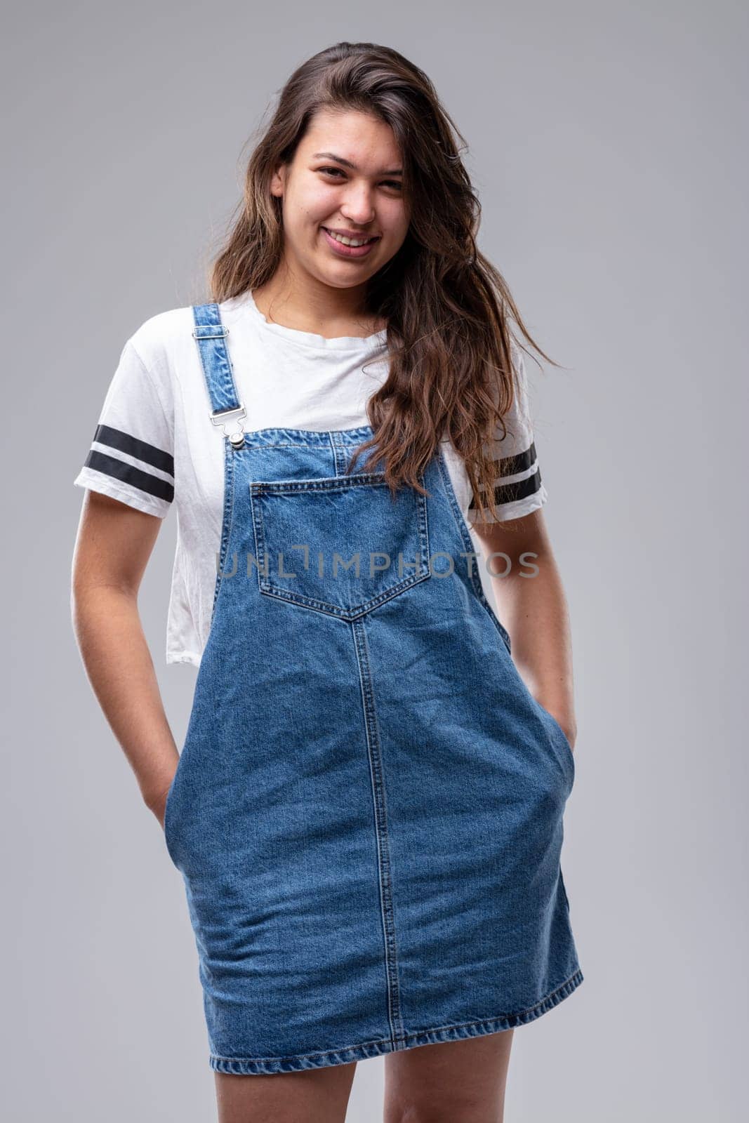 Confident casual trendy young woman in a denim pinafore standing in a relaxed pose with hands in pockets giving the camera a lovely friendly smile