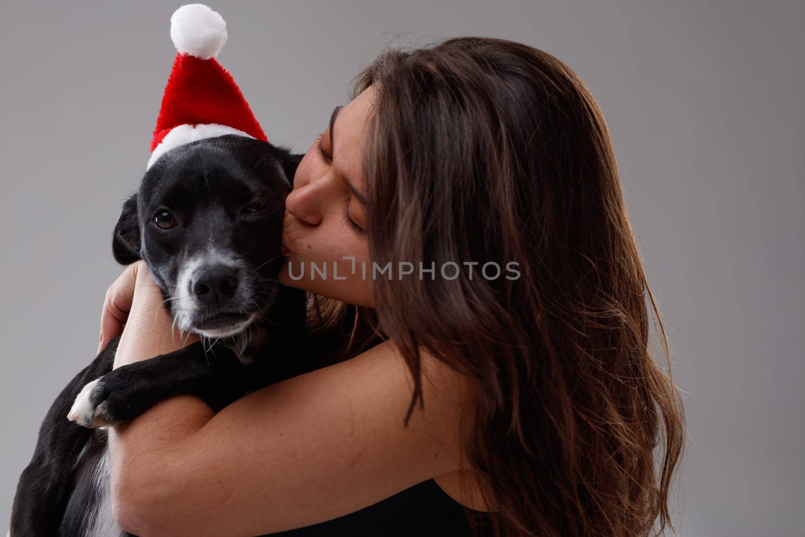 Cute little dog wearing a red Father Christmas hat being kissed by its loving young female owner in a close up portrait celebrating the holiday season