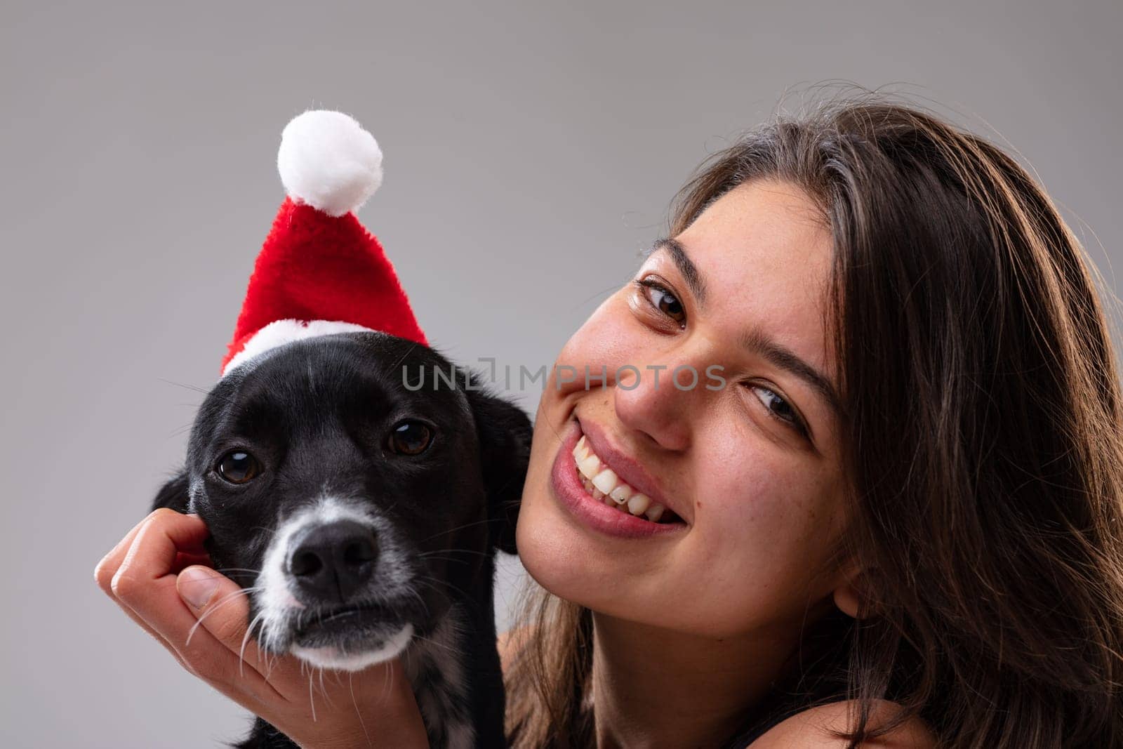 Cute little dog wearing a Santa hat with a smiling young woman alongside to celebrate the Christmas holiday season in a close up on their faces