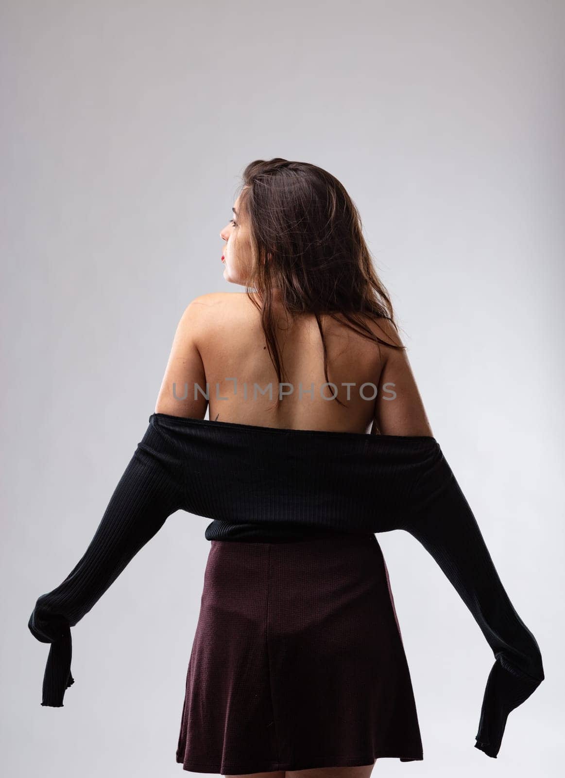 Trendy young woman posing with her black pulled top down exposing her bare her shoulders in a three-quarter back view on grey