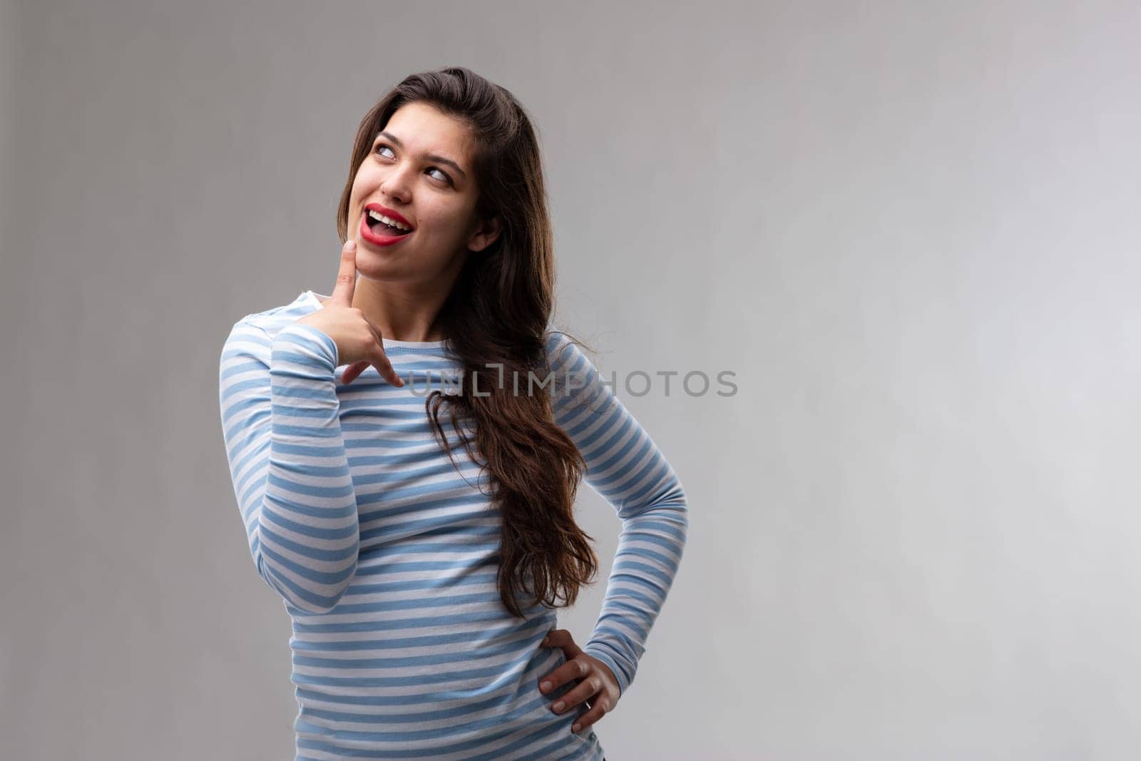 Thoughtful young woman with a look of excited anticipation standing with a finger to her open mouth in deep concentration as she lets her imagination run free over a grey studio background