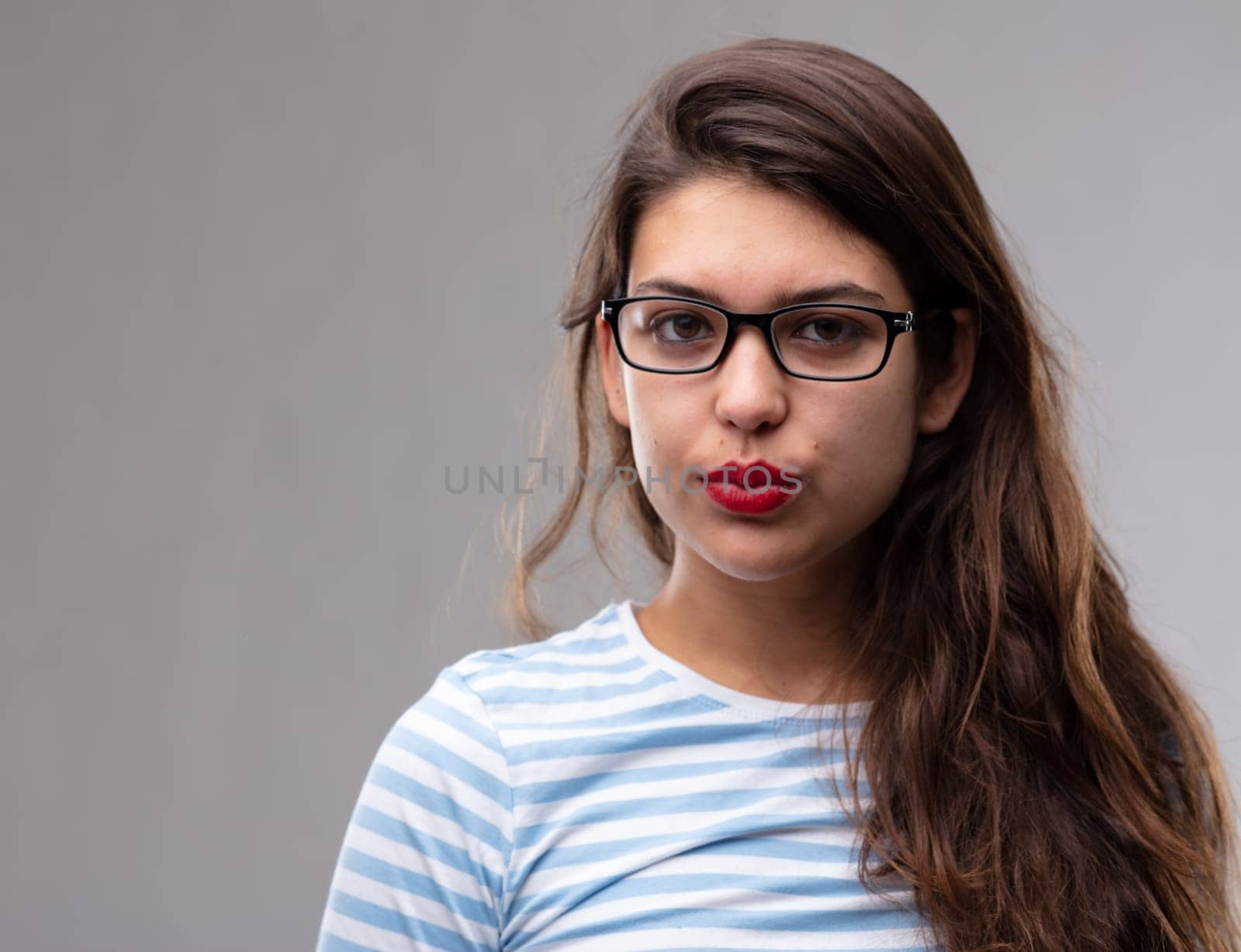 Young dark haired woman wearing glasses staring intently at the camera with a thoughtful expression pouting her red lips over a grey studio background
