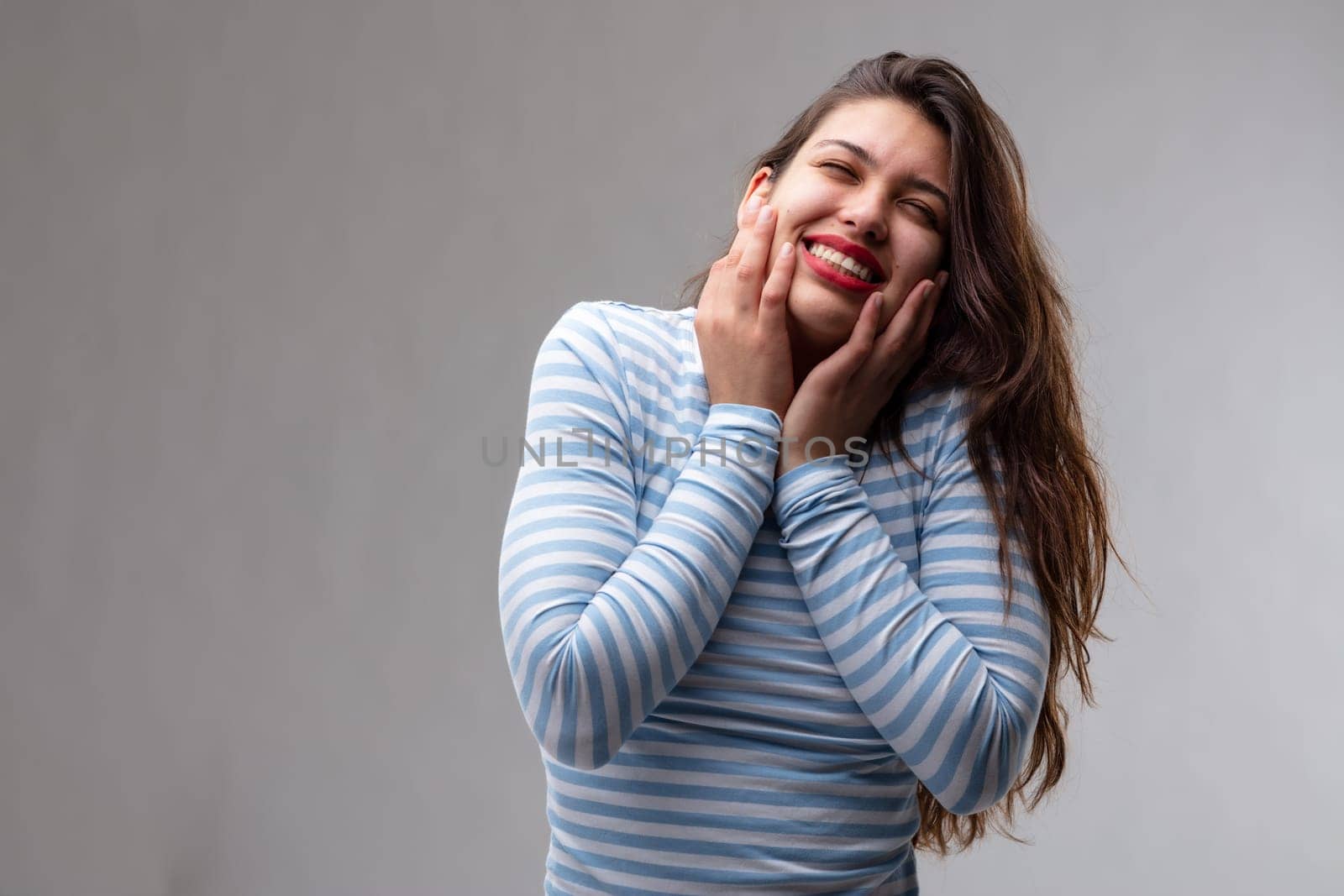 Happy relaxed friendly young woman with a beaming smile and hands to her cheeks tilting her head to the side over a grey studio background with copy space