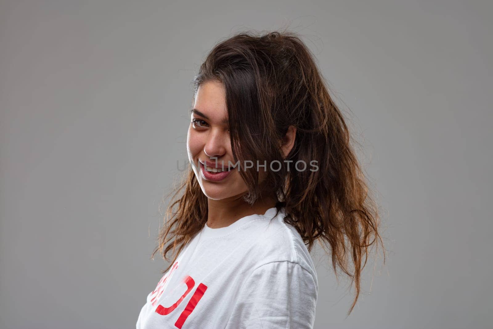 Cute attractive young woman with mussed up tousled long hair turning to look back at the camera with a lovely friendly smile in a close up studio portrait