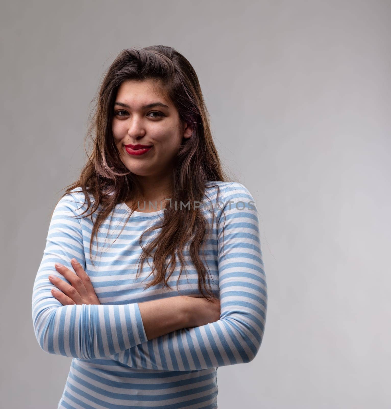Happy attractive confident young woman with an amused smile standing with folded arms looking at the camera over a grey studio background