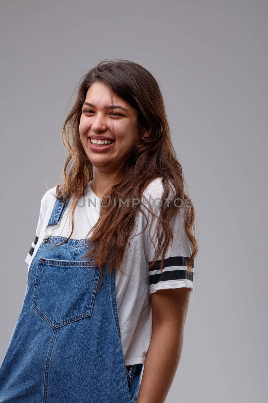 Laughing happy young woman in trendy denim dungarees standing with hands in pockets and a vivacious smile over a grey wall