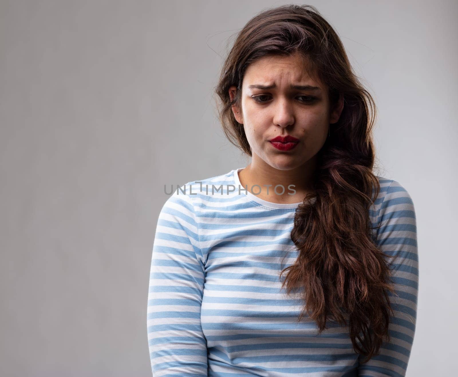 Anxious worried moody young woman puckering up her face and frowning as she looks down with a sad expression over a grey studio background