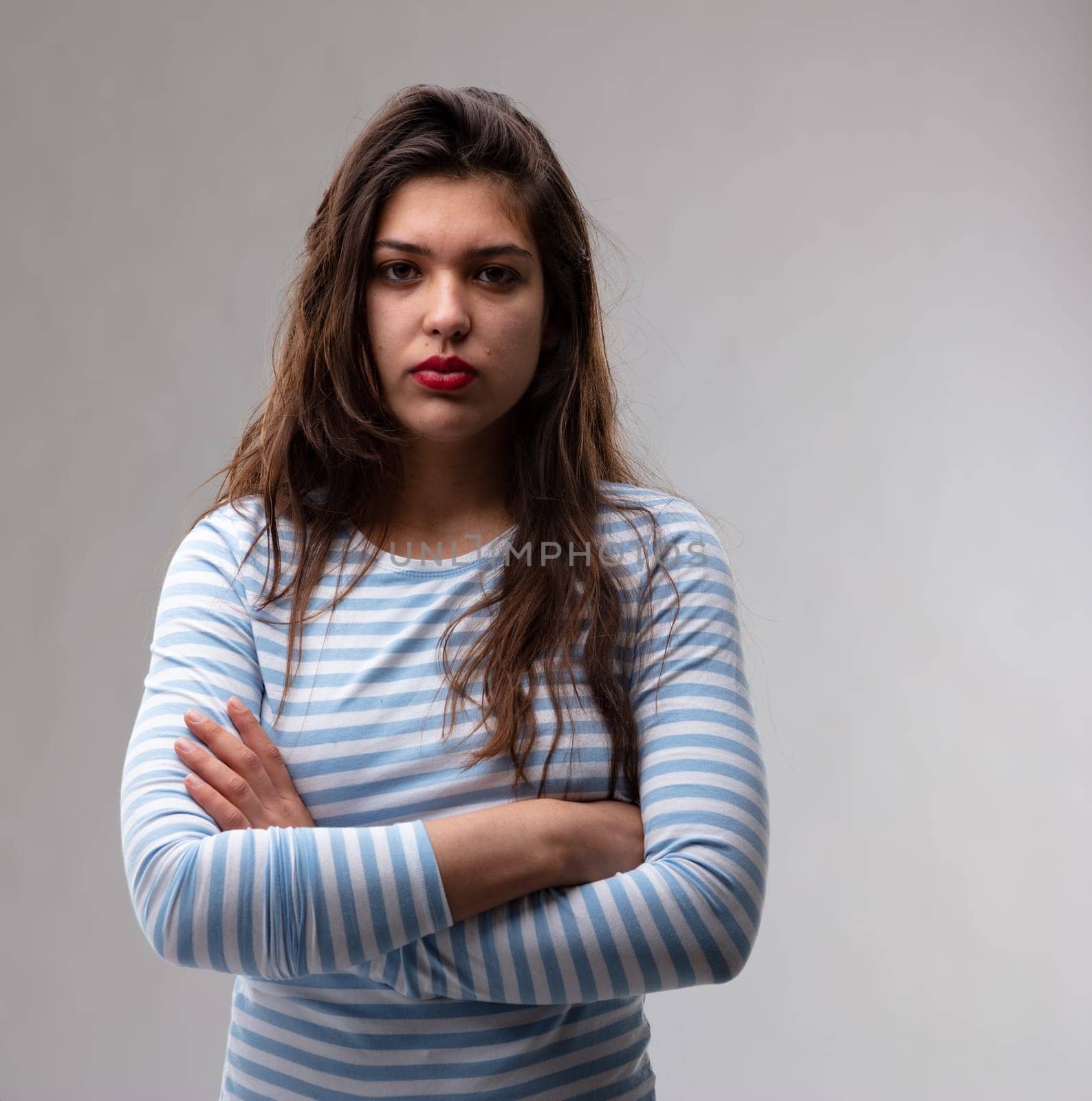Young woman with a deadpan stern expression and penetrating stare standing with folded arms looking at the camera over a grey studio background