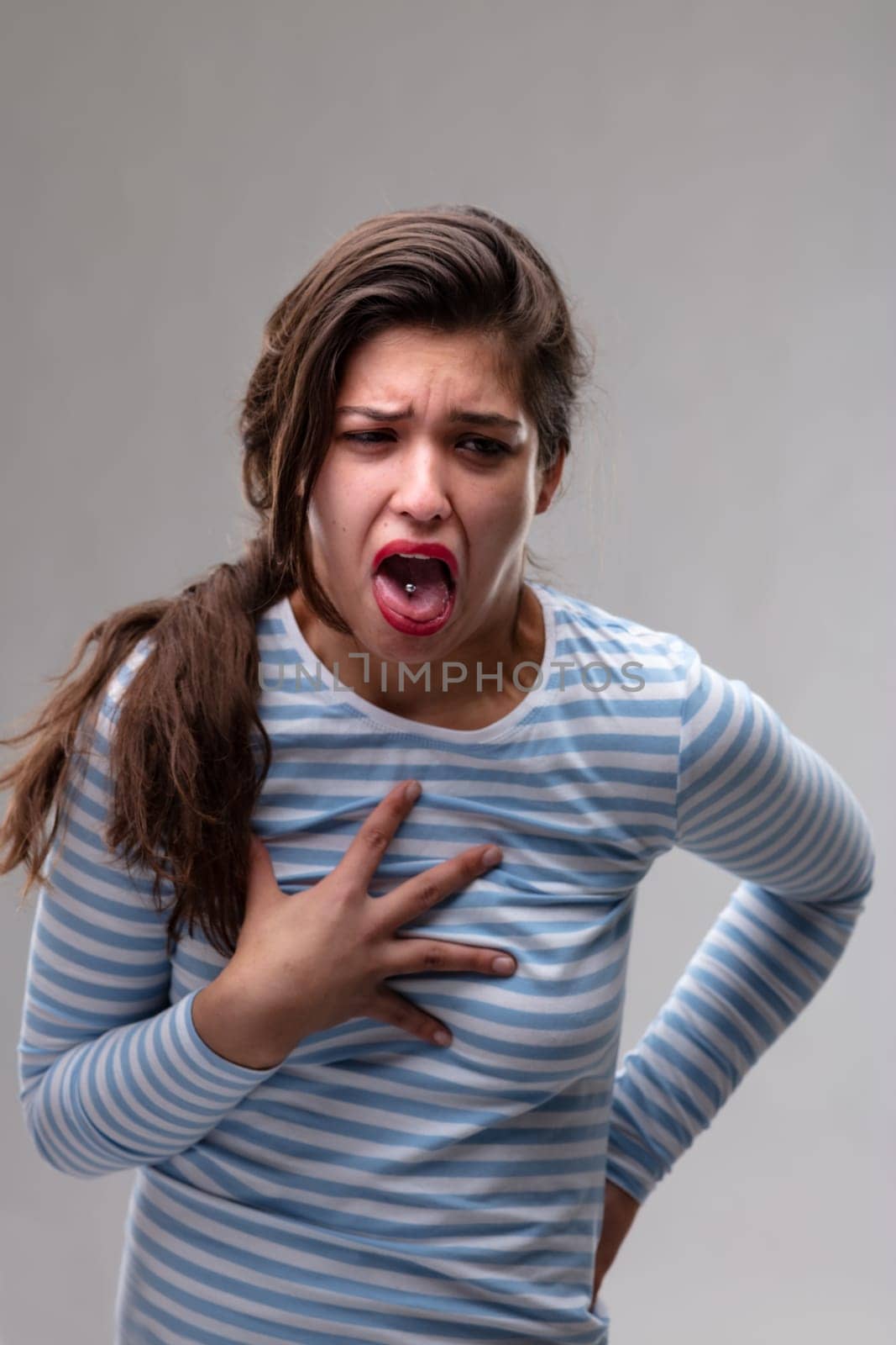 Young woman expressing loathing or pain clutching her chest and back grimacing and making a vomiting gesture with tongue out over a grey background