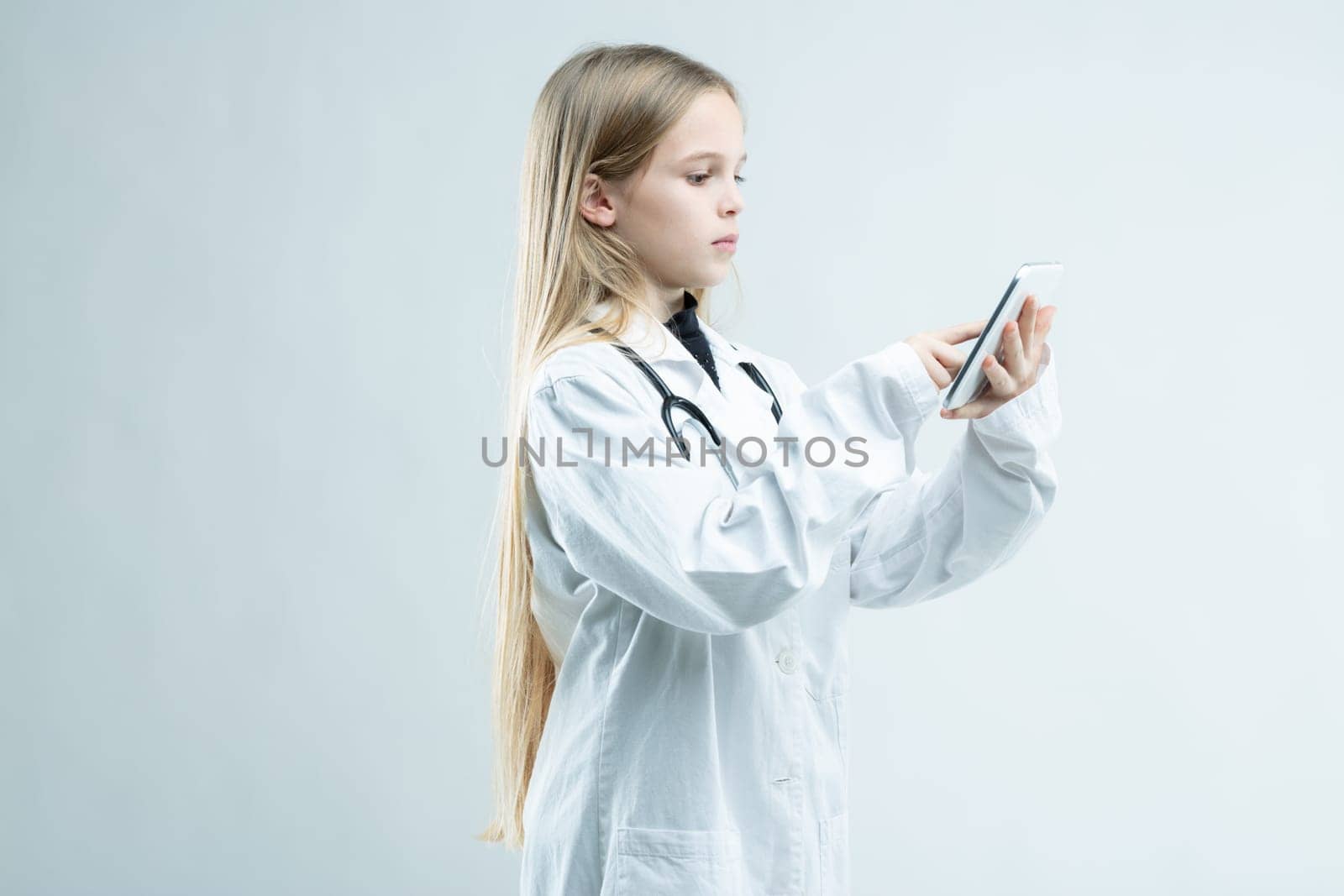 Young girl dressed up in a medical lab coat with stethoscope pretending to be a doctor standing using her mobile phone isolated on white