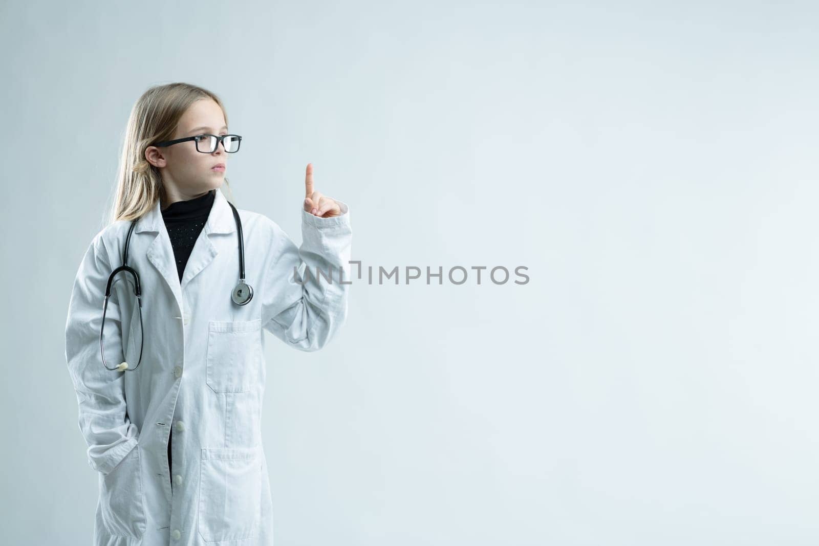 Cute little blond girl playing at being a doctor in lab coat and stethoscope holding up her finger looking towards blank copy space on white