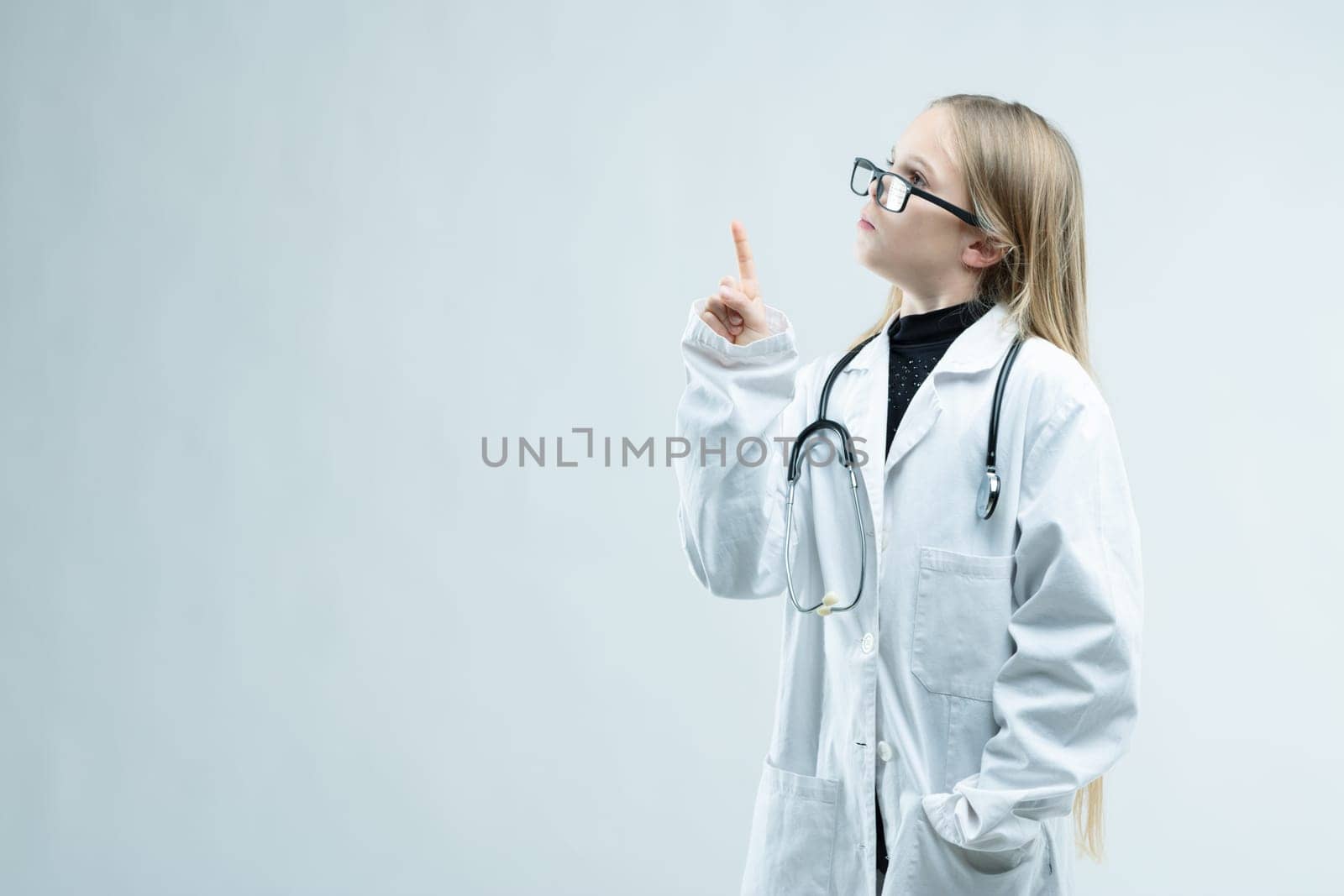 Little girl dressed up as a nurse or doctor in outsize glasses, white coat and stethoscope holding up her finger towards blank copy space on white
