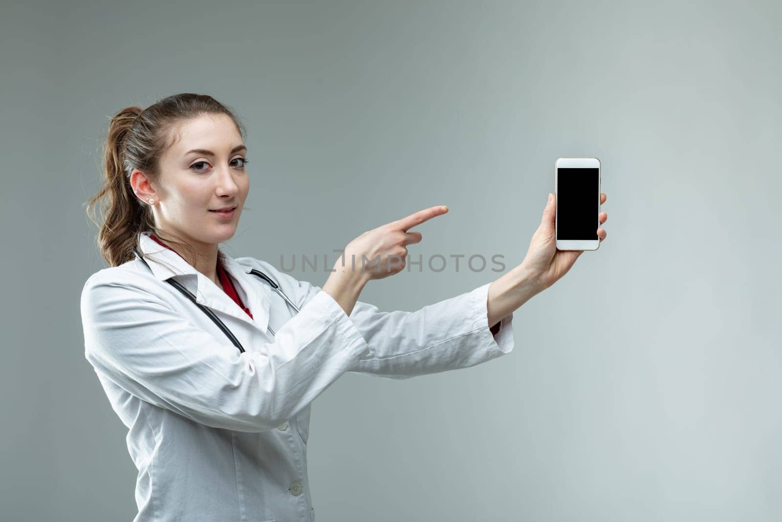Serious doctor pointing to her mobile phone as she holds it extended in her hand with the blank screen to the viewer over grey with copy space