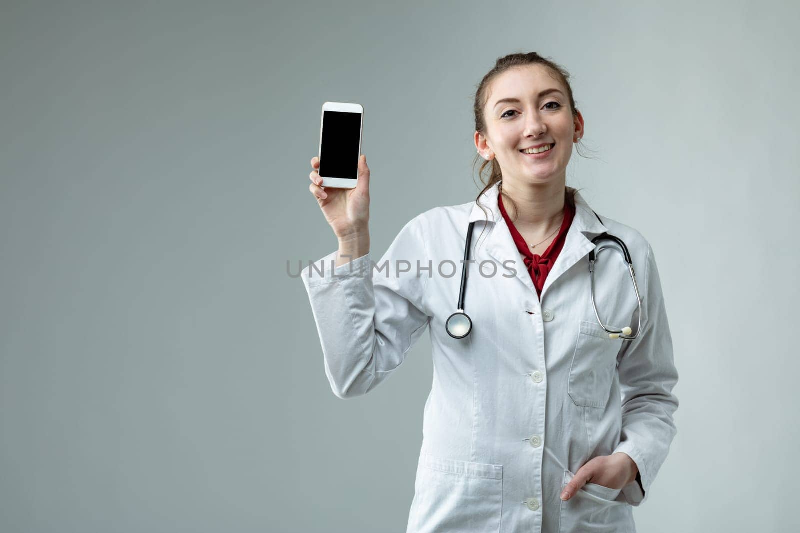 Smiling woman doctor in white lab coat and stethoscope holding up a mobile phone with the blank screen to the camera with a happy smile over grey with copy space