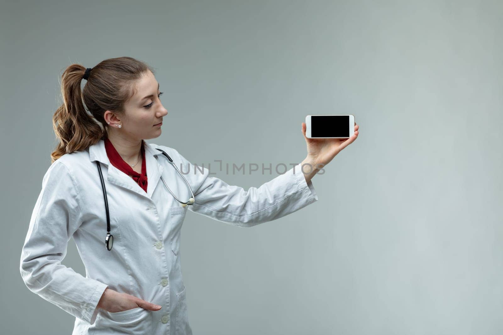 Woman doctor or nurse wearing a stethoscope and white lab coat standing holding up her mobile phone with the screen to the camera over grey wth copy space
