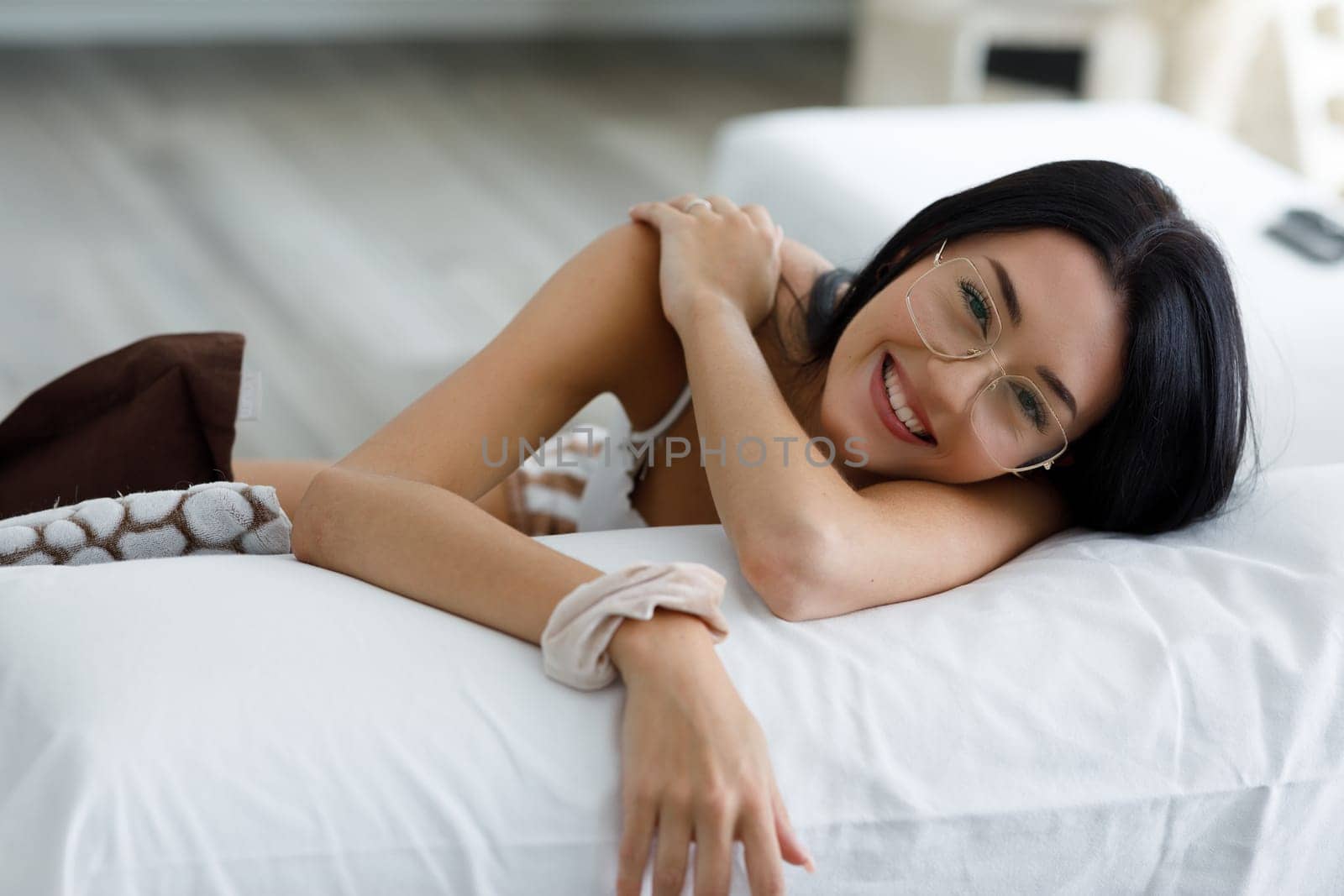 Pretty vivacious young woman wearing gold rimmed glasses relaxing on a sofa looking over the back at the camera with a happy friendly smile in close up