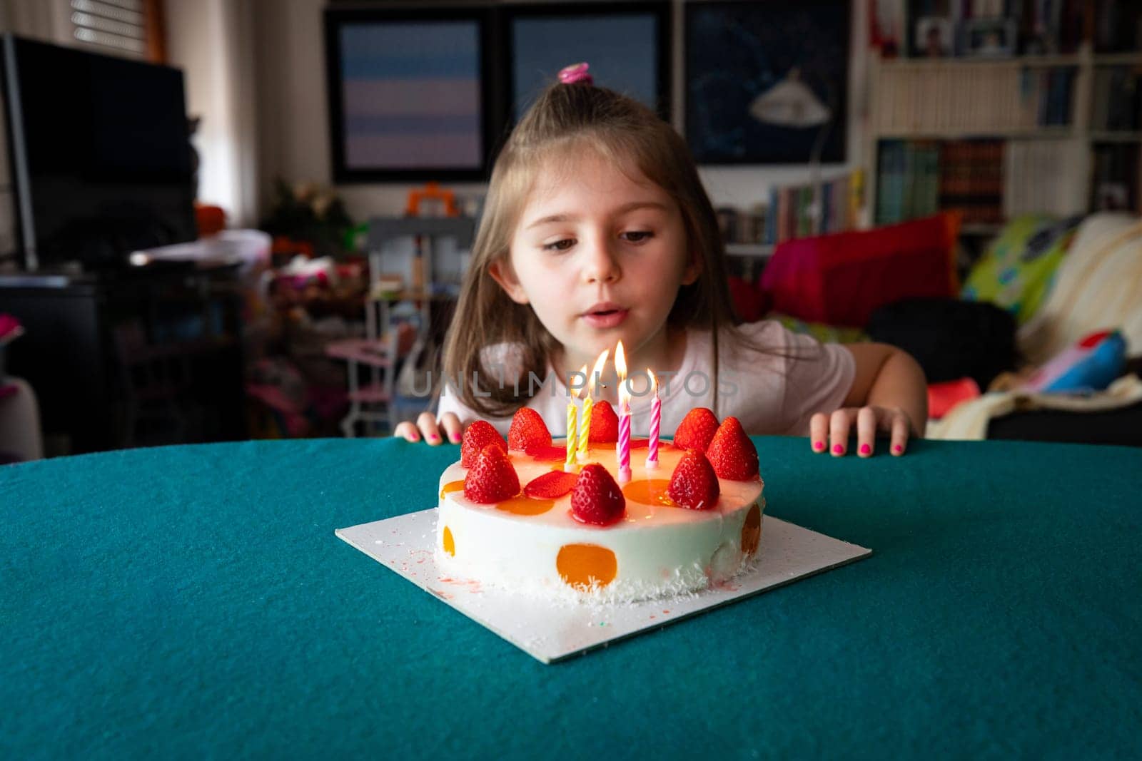 Adorable little girl blowing out five candles on her birthday cake topped with fresh ripe strawberries at home in the living room with copy space in a close up portrait