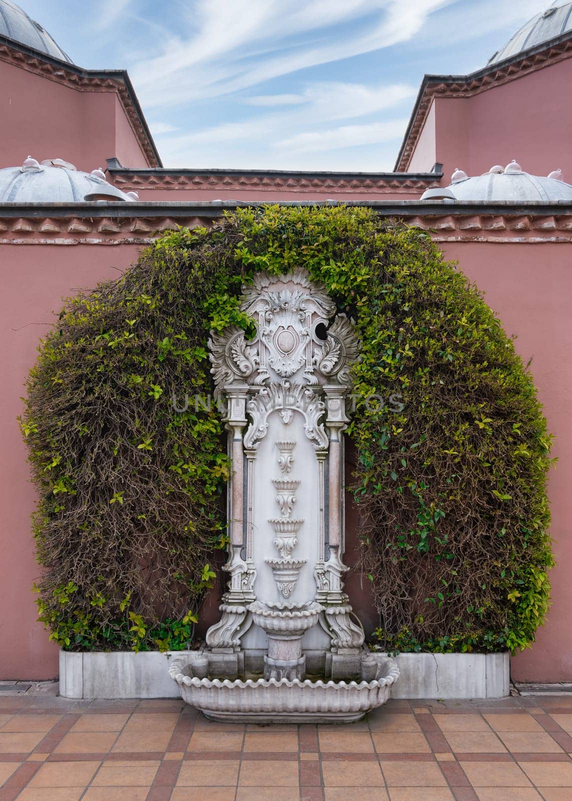 White marble fountain in Sultan Ahmed Square, Istanbul, Turkey, surrounded by lush greenery and a pink stone wall of Hagia Sophia Hurrem Sultan Bathhouse, or Ayasofya Hurrem Sultan Hamami