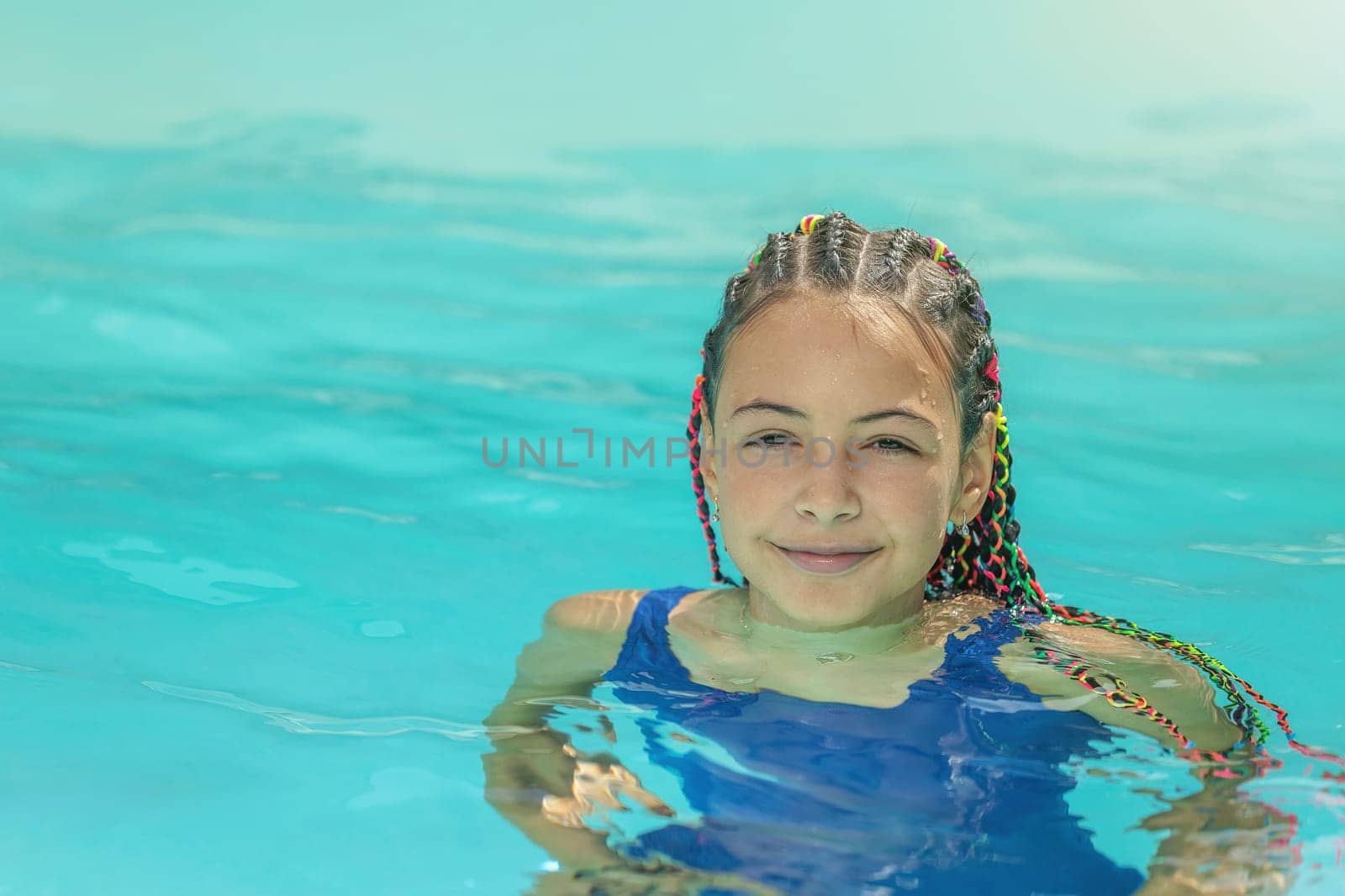 Little smiling long haired girl is in a swimsuit standing in the pool outside .Horizontally.