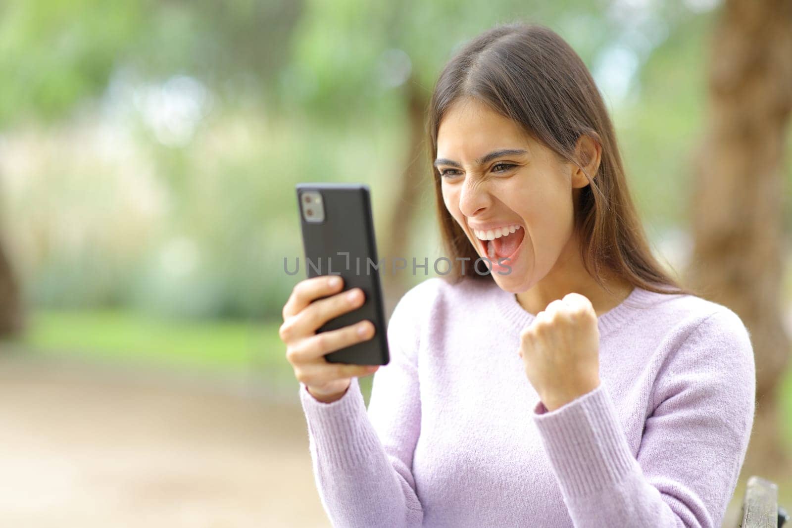 Excited woman celebrating good news watching phone content in a park