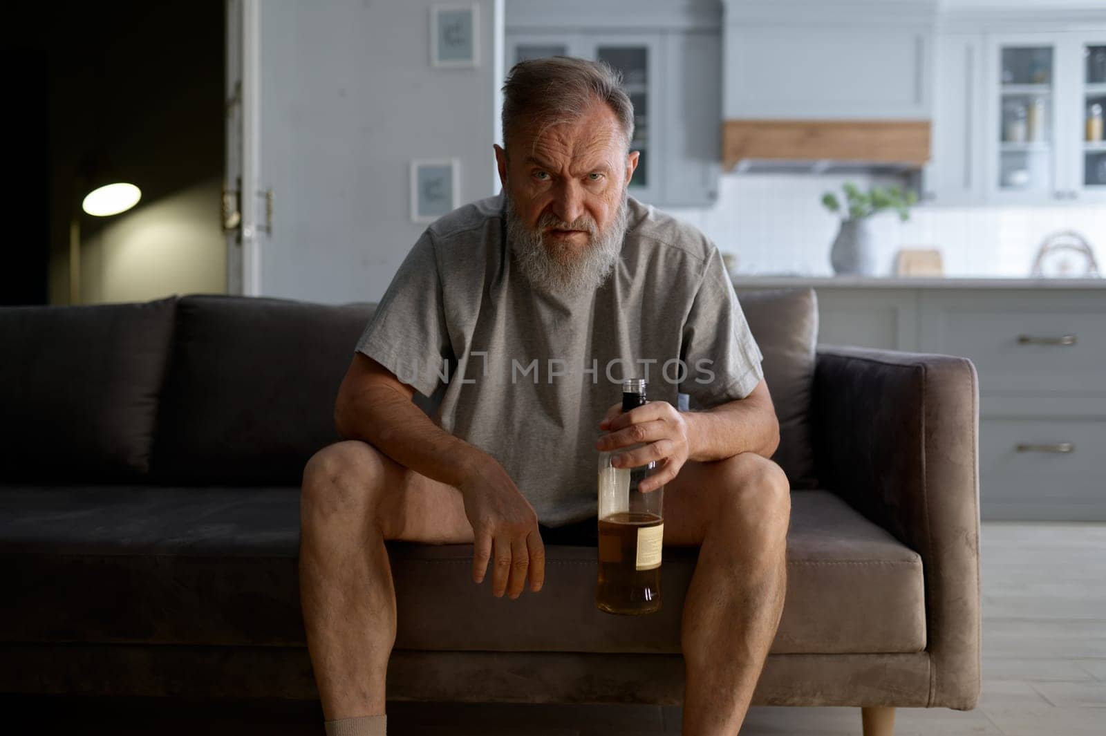 Drunk senior man looking at camera with angry rage emotion on face while sitting on sofa at home living room. Old veteran and ptsd problem. Alcohol addiction and retirement