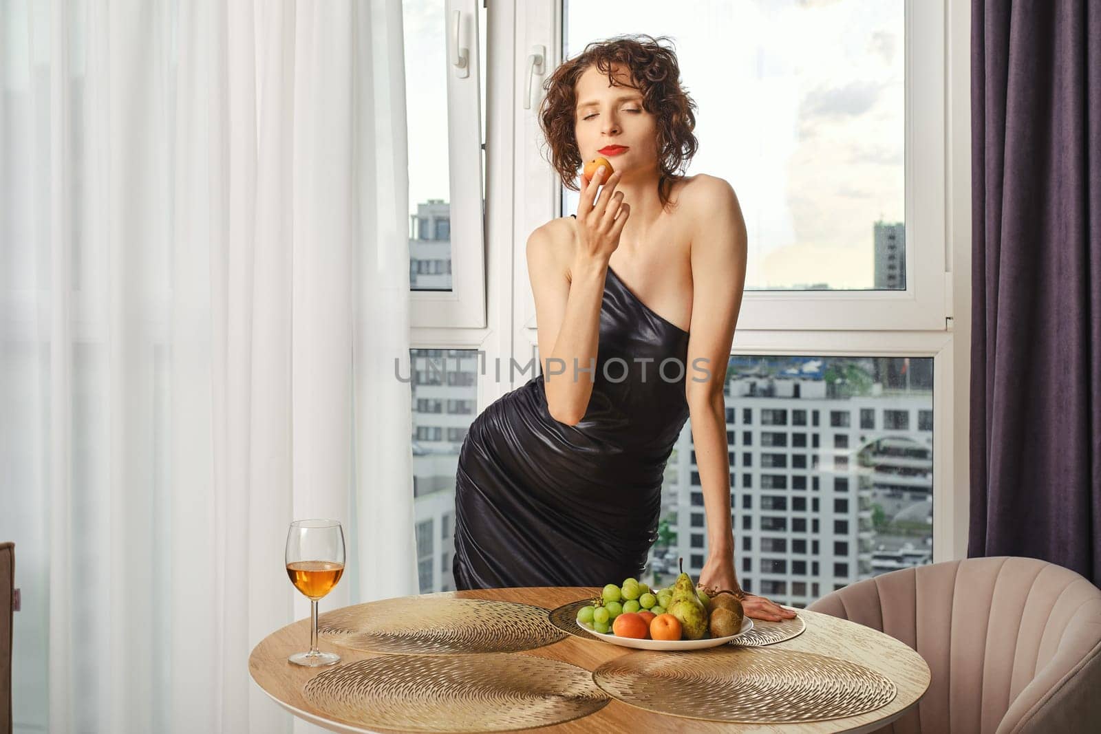 Stylish woman ready for party biting apricot before leaving apartment
