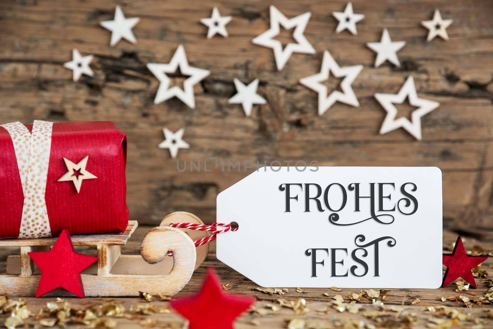 Wooden Sled With Red Gift And Rustic Christmas Background With Label With German Text Frohes Fest, Which Means Happy Holidays In English
