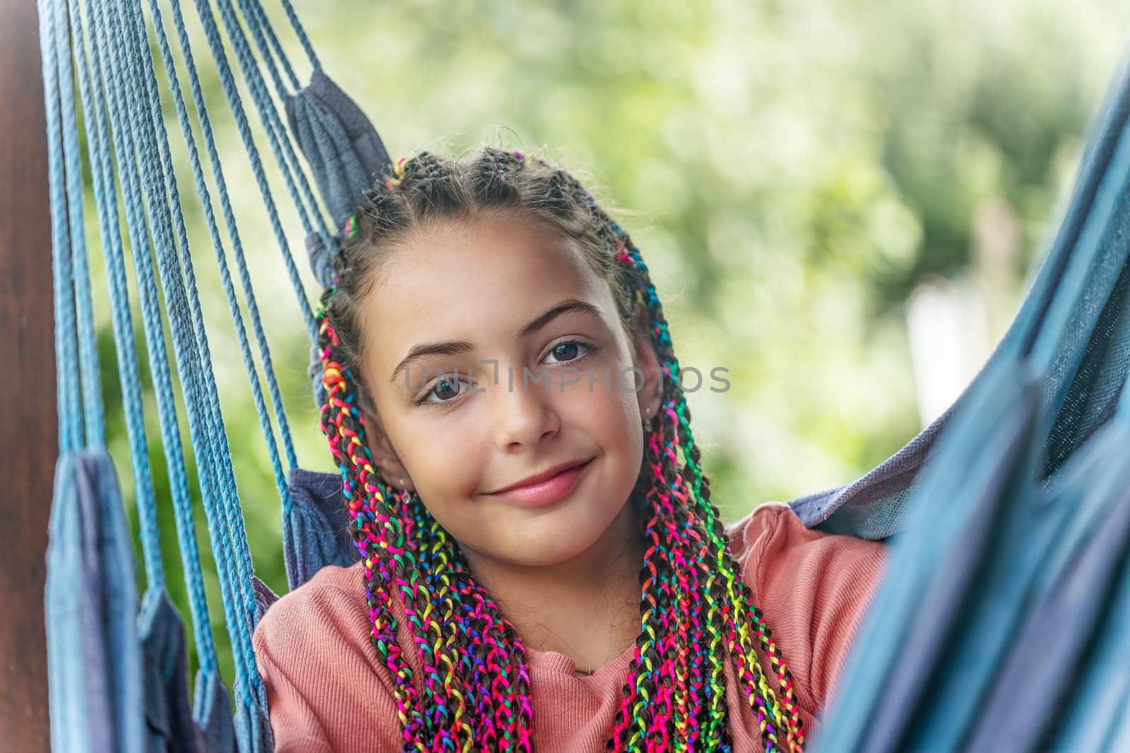 Smiling young girl with colorful braids in her hair is posing on blue hammock. Horizontally.