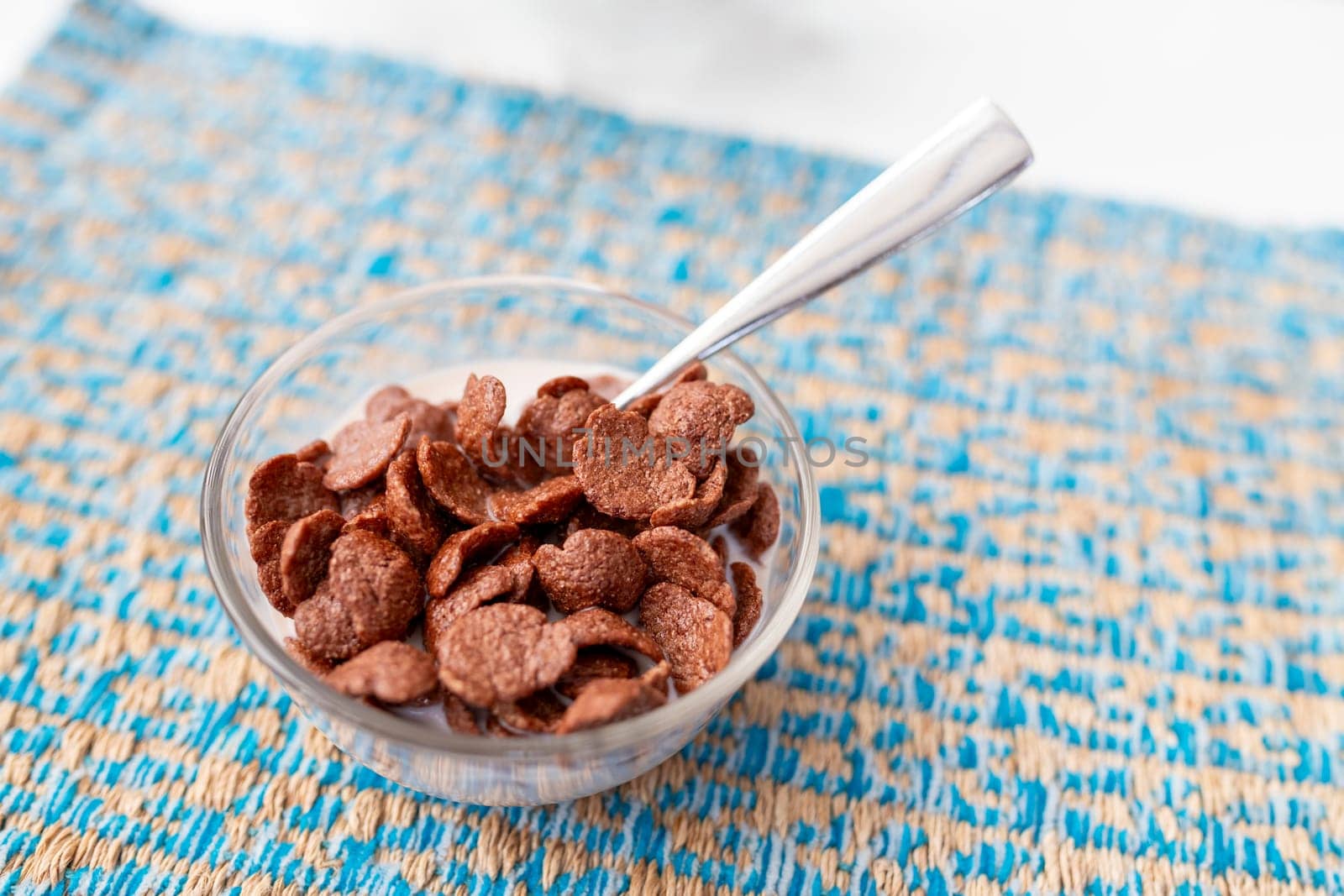 Shallow focus shot of chocolate-flavored breakfast cereal in a clear, circular glass dish on a patterned blue saucer, complete with stainless steel spoons. On a table in a hotel restaurant.