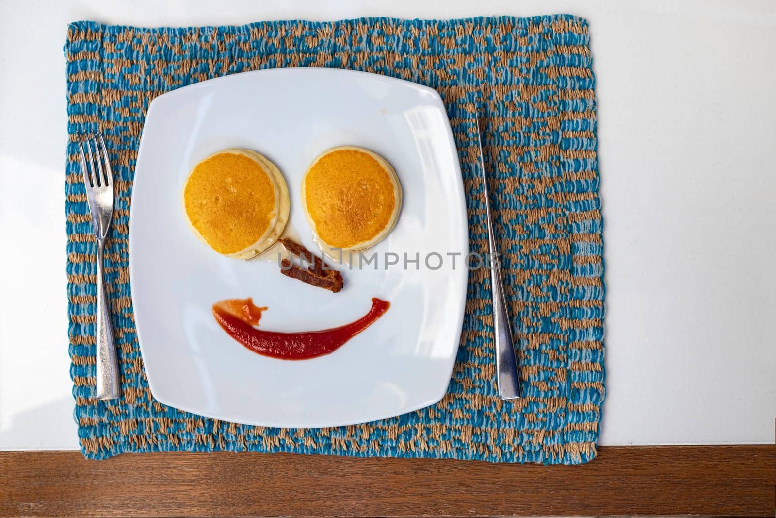 Top view of fork and knife next to breakfast plate on dining table On the plate were pancakes, syrup, crispy bacon and ketchup. arranged into smiley faces Refers to waking up to work brightly.