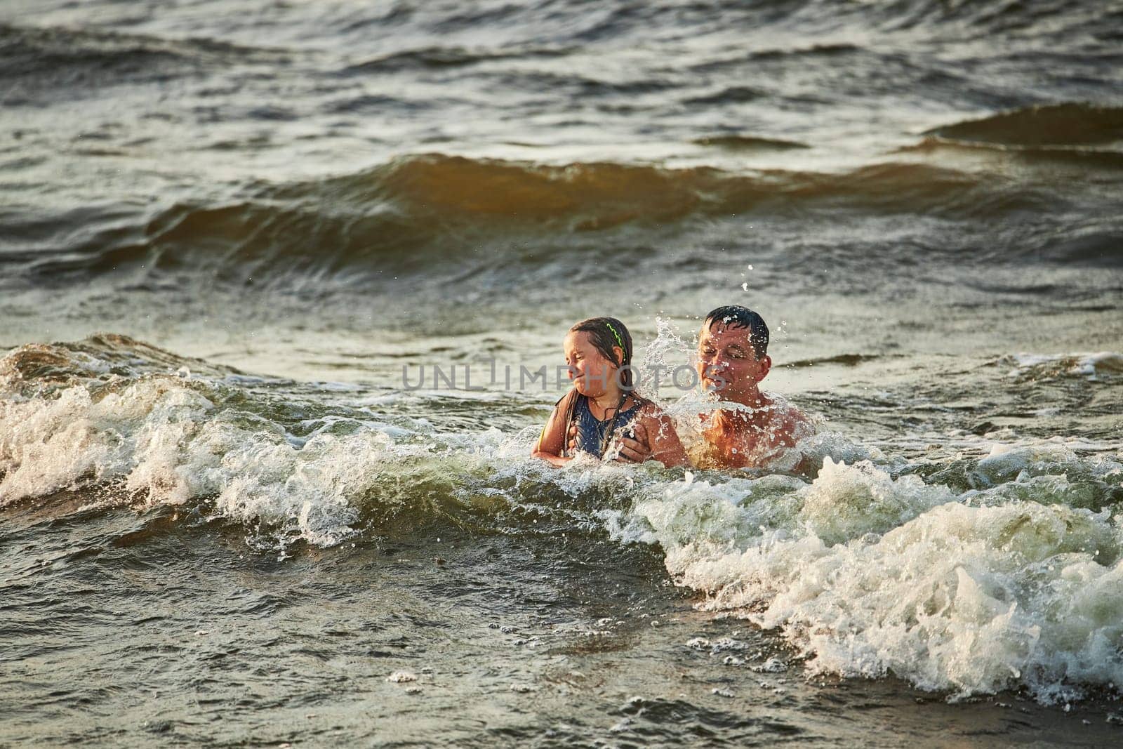 Little girl and her dad playing with waves in the sea. Family summer vacations. Kid playfully splashing with waves together with father. Jumping in sea waves. Summer vacation on the beach. Water splashes. Travel during summertime concept