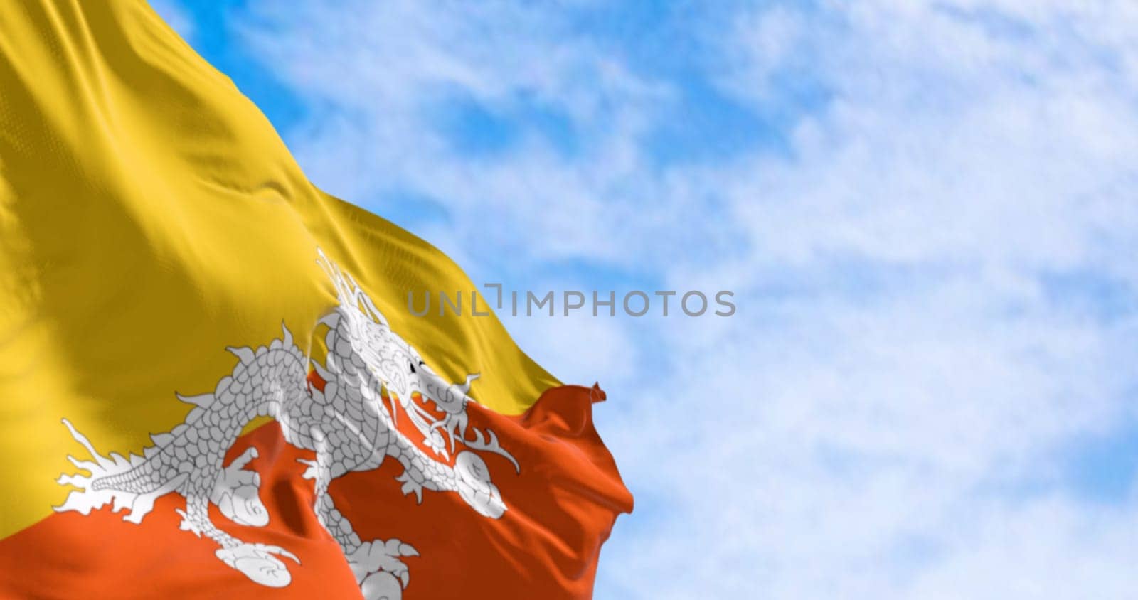 National flag of Bhutan waving in the wind on a clear day. Yellow upper triangle and an orange lower triangle, featuring a white dragon holding jewels in the center