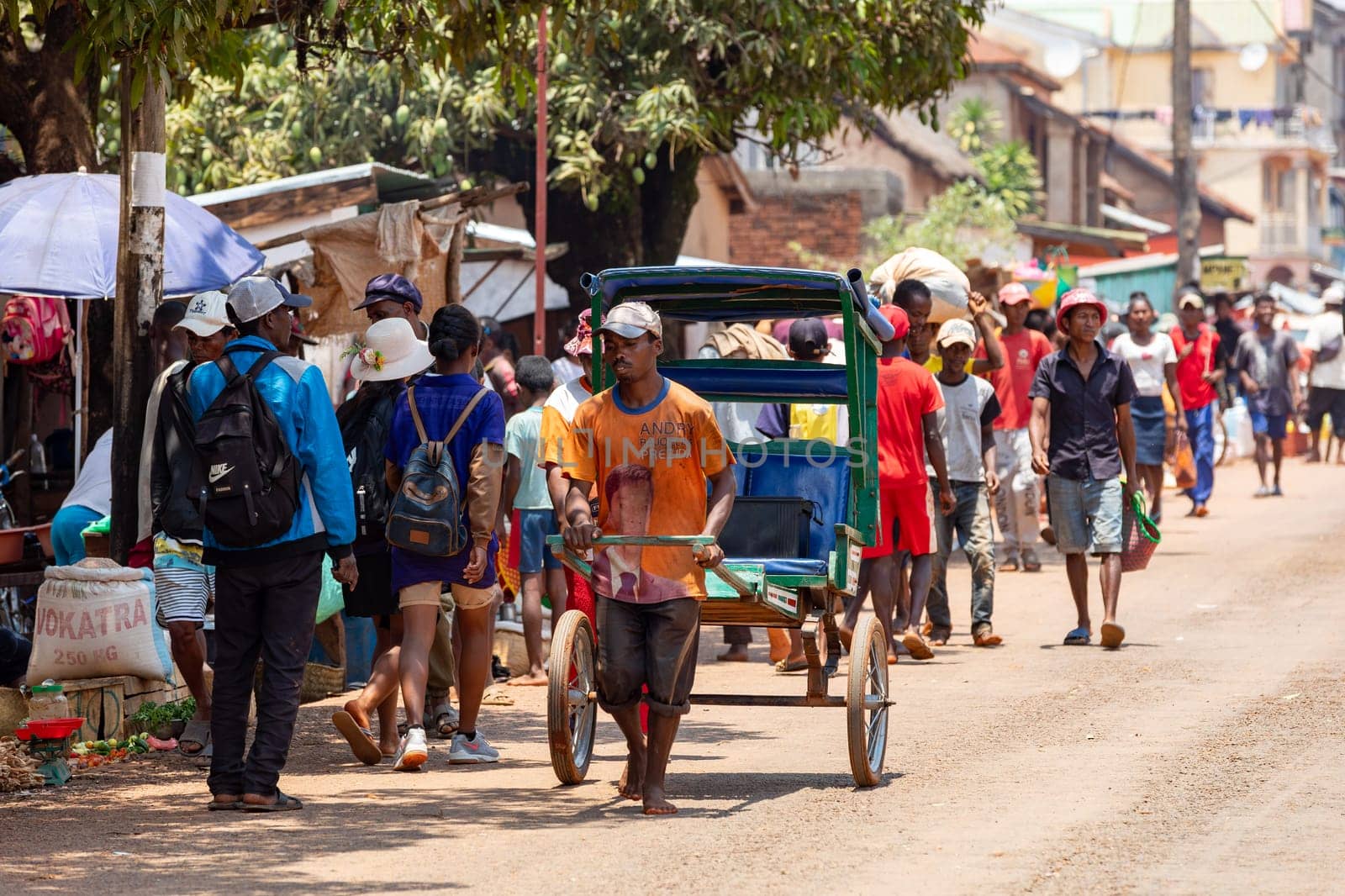Mandoto, Madagascar - November 2nd 2022: Rickshaw runner dashes through the crowded streets with small wooden cart. A handheld rickshaw driven by a running person is traditional mode of transport.