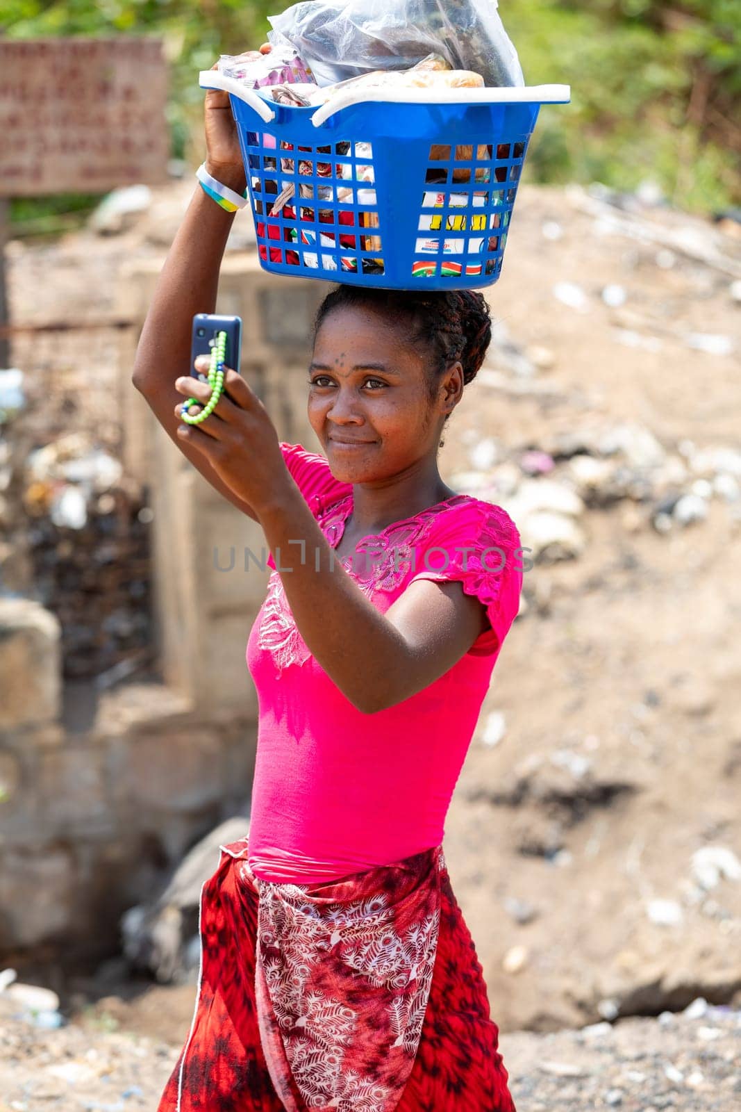Miandrivazo, Madagascar - November 2nd, 2022: A woman carrying a basket on her head captures a photo using an old-fashioned mobile phone.