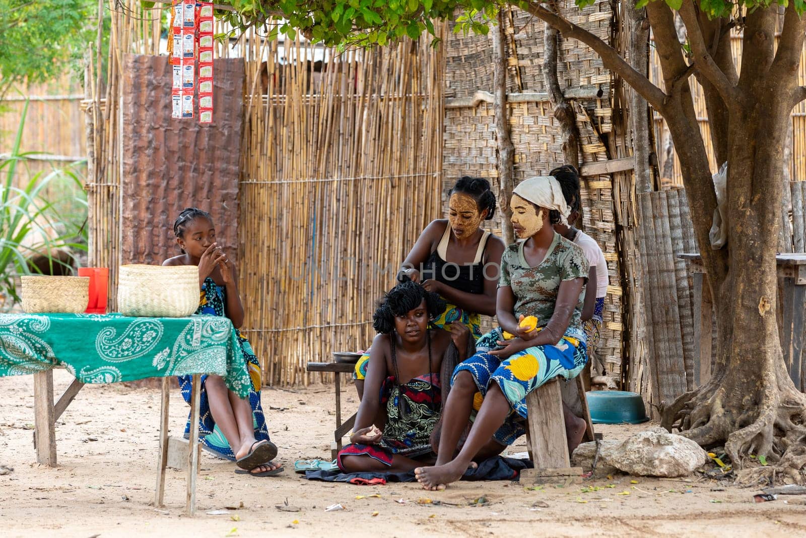 Bekopaka Ferry Port, Madagascar - November 4th 2022: Face painted Malagasy woman styling hair to other woman in front of her hut.