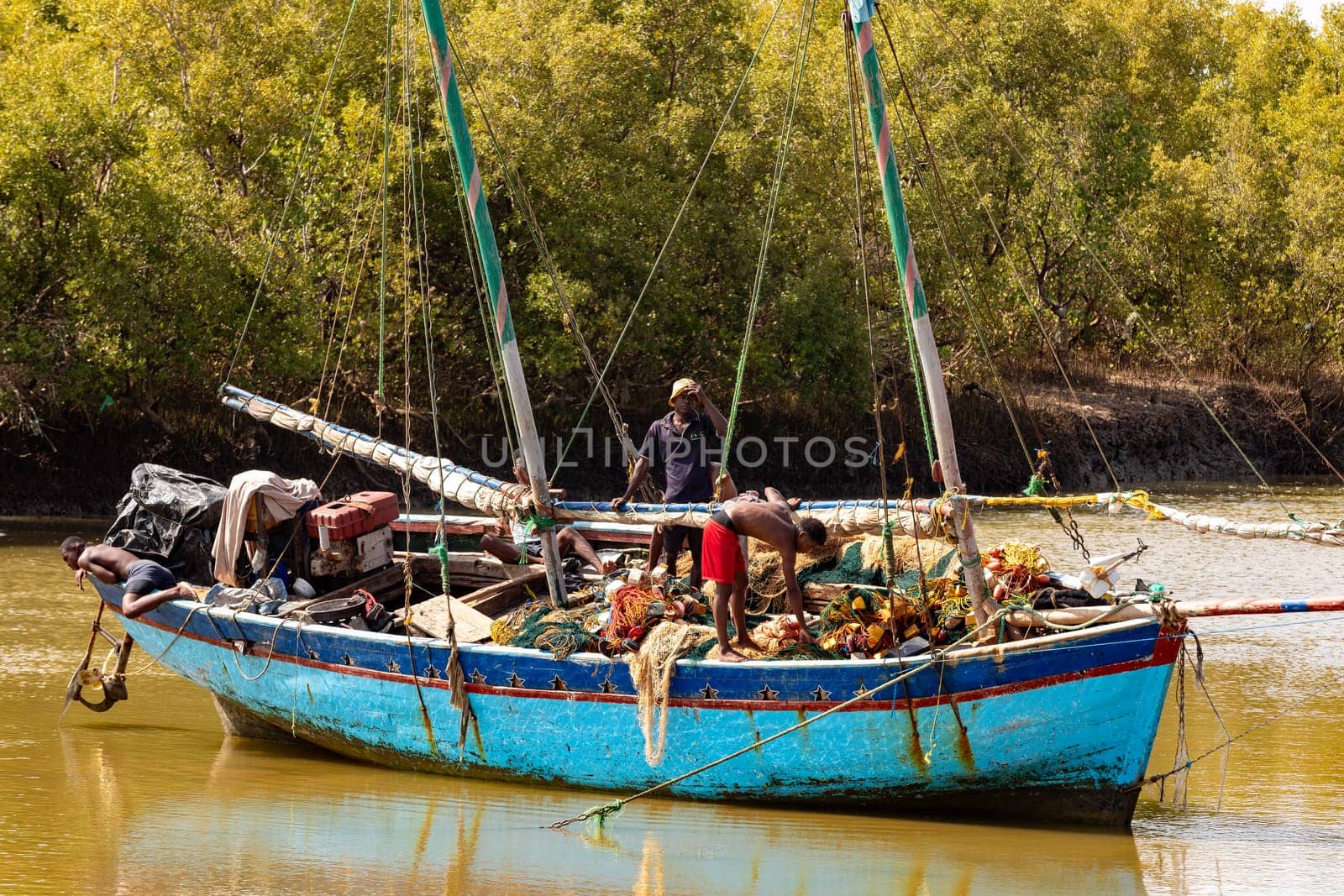 MORONDAVA, MADAGASCAR - AUGUST 28 2023: Two men repair a fishing net on a blue boat moored to the riverbank in Morondava. The boat is equipped with two masts and other fishing gear.