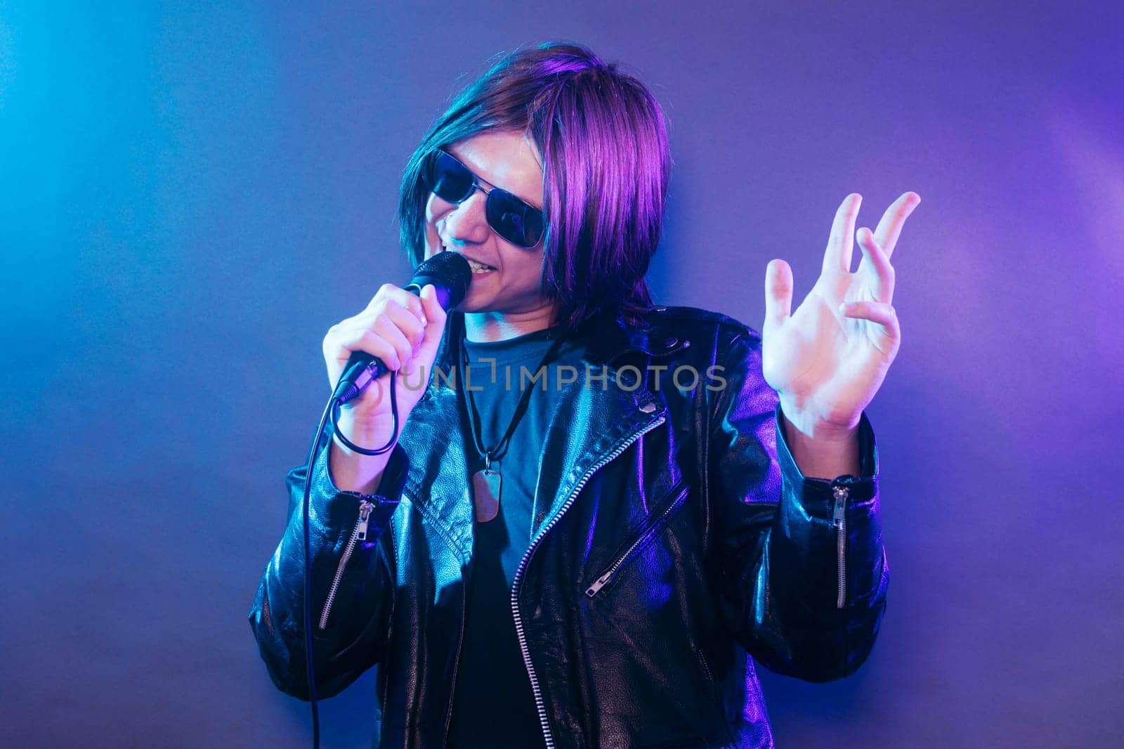 The young rock star in black leather is singing in microphone on blue purple background.