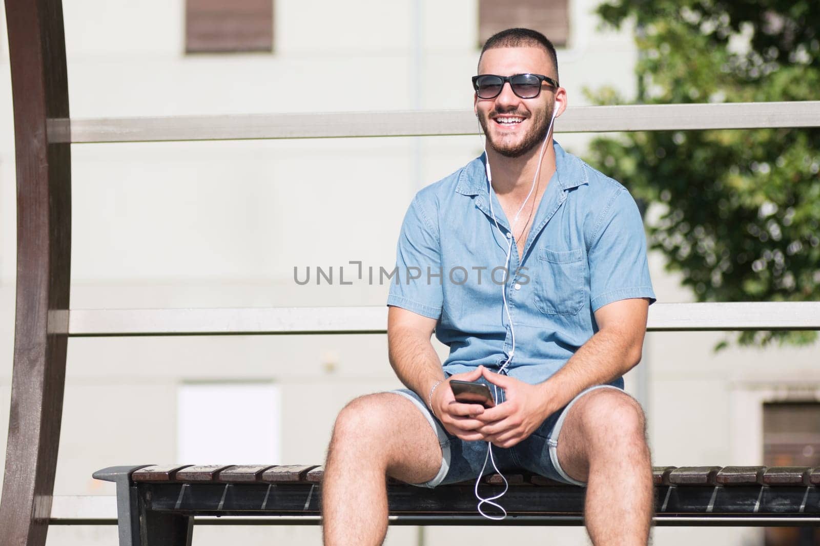 Smiling young guy with earphones is sitting on bench and using phone.